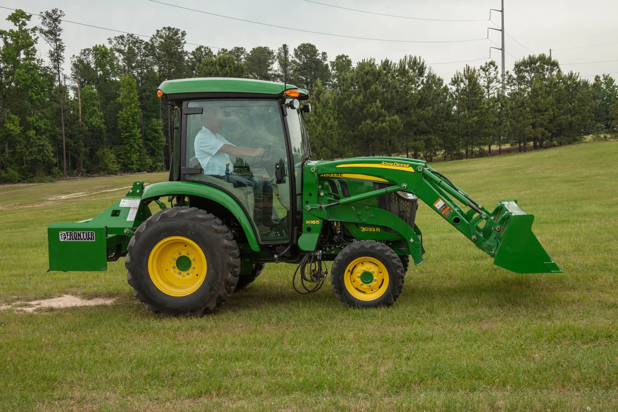 Side view of green tractor with front loader attachment on grass, with an operator inside the cab and trees lining the background under an overcast sky