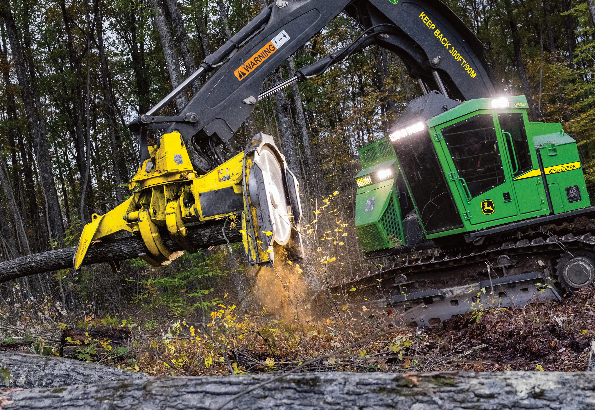 John Deere tracked forestry machine cuts a tree with a harvesting head, sending wood chips outward, in a dense forest with fallen logs and debris covering the ground.