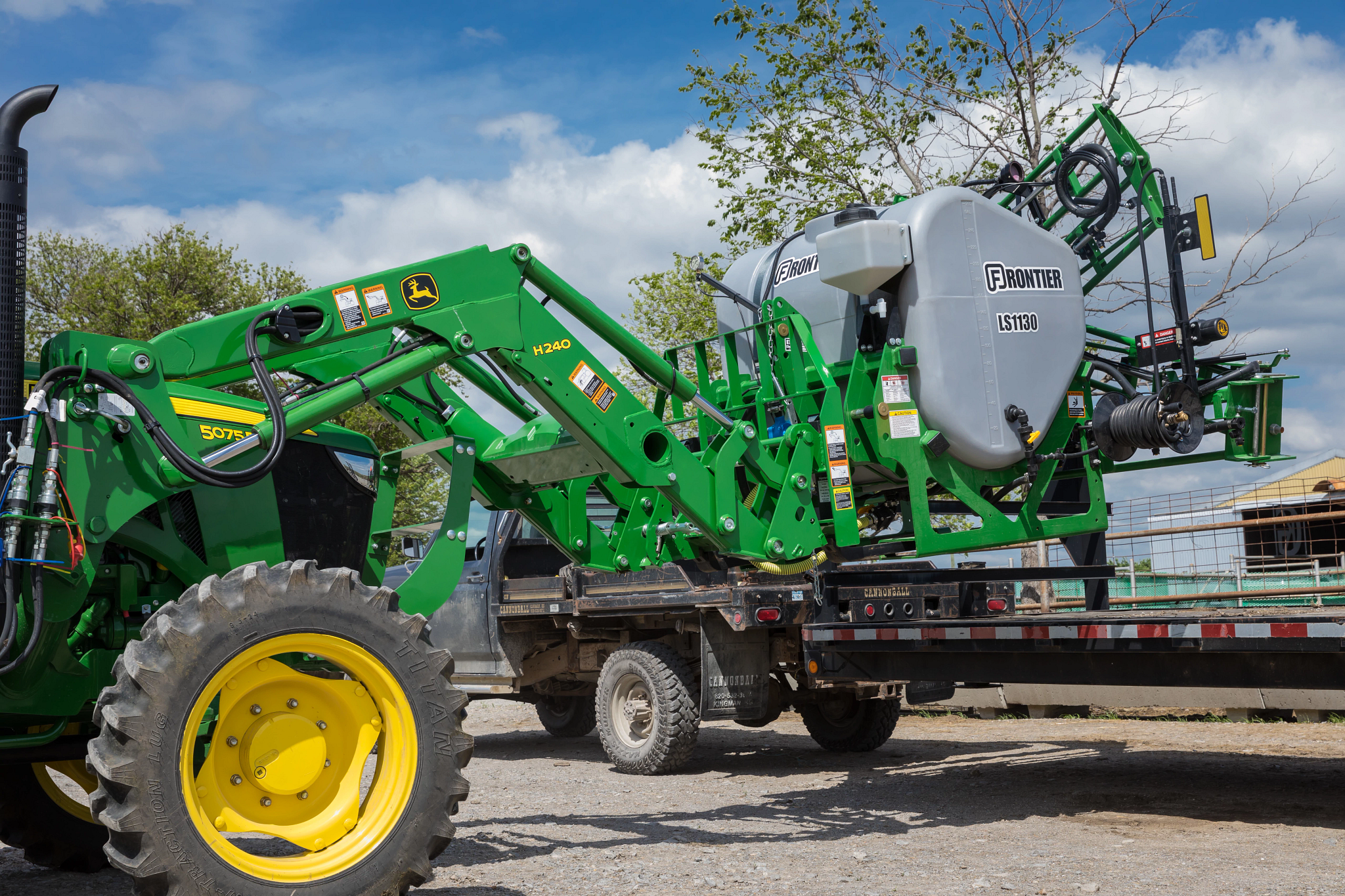 John Deere tractor with a front loader lifting and loading a large sprayer tank labeled onto a flatbed truck outdoors under a partly cloudy sky.