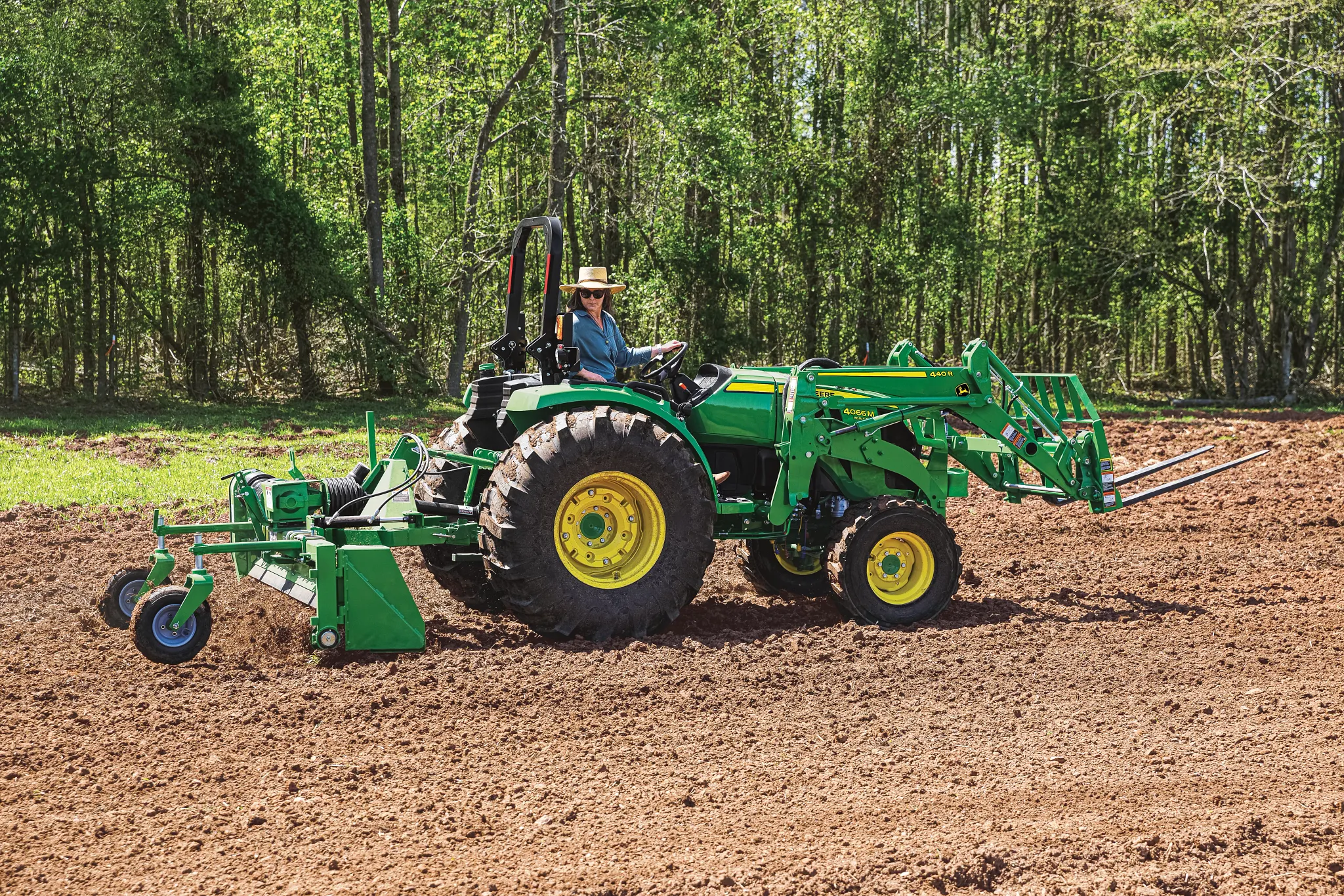 Tractor with a front loader and tilling attachment drives across a brown, freshly tilled field. A person in a hat operates the tractor, with a line of leafy trees in the background under bright daylight.