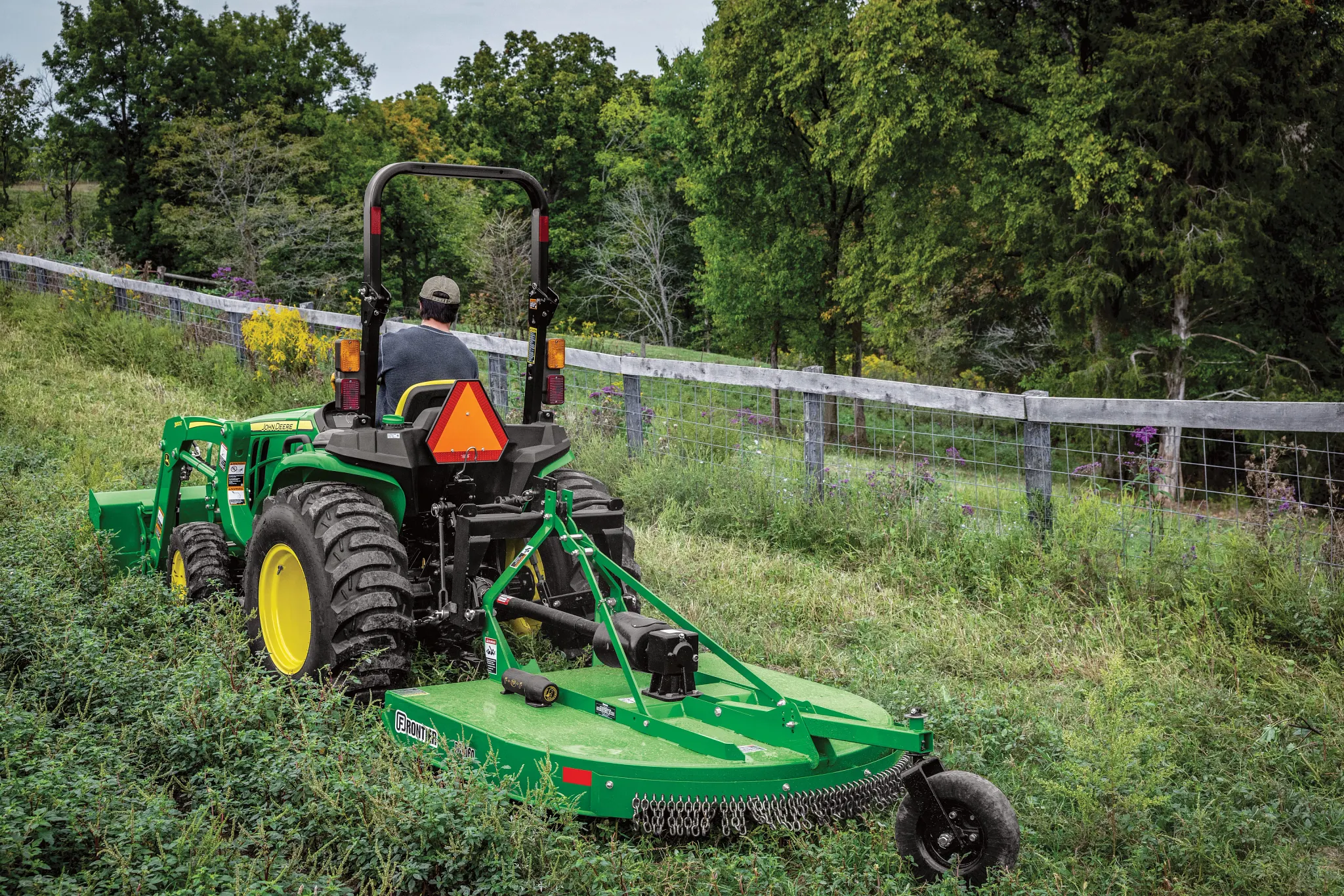 An operator on a green tractor with a rear-mounted mower cuts grass in a sloped field, with a wire fence and dense trees lining the background in a rural setting
