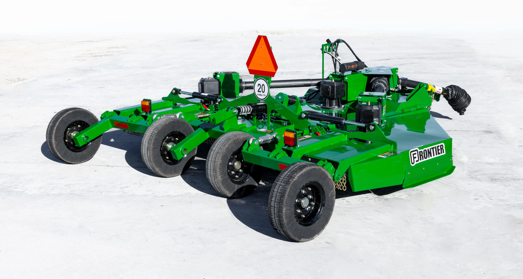 Rear three-quarter view of a green Frontier rotary cutter in transport mode, featuring four transport wheels, PTO driveline, hydraulic components, warning lights, safety chain shielding, and a slow-moving vehicle emblem on a concrete surface