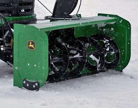 Front view of a snow blower attachment with black spiral auger blades clearing snow on a snowy surface.