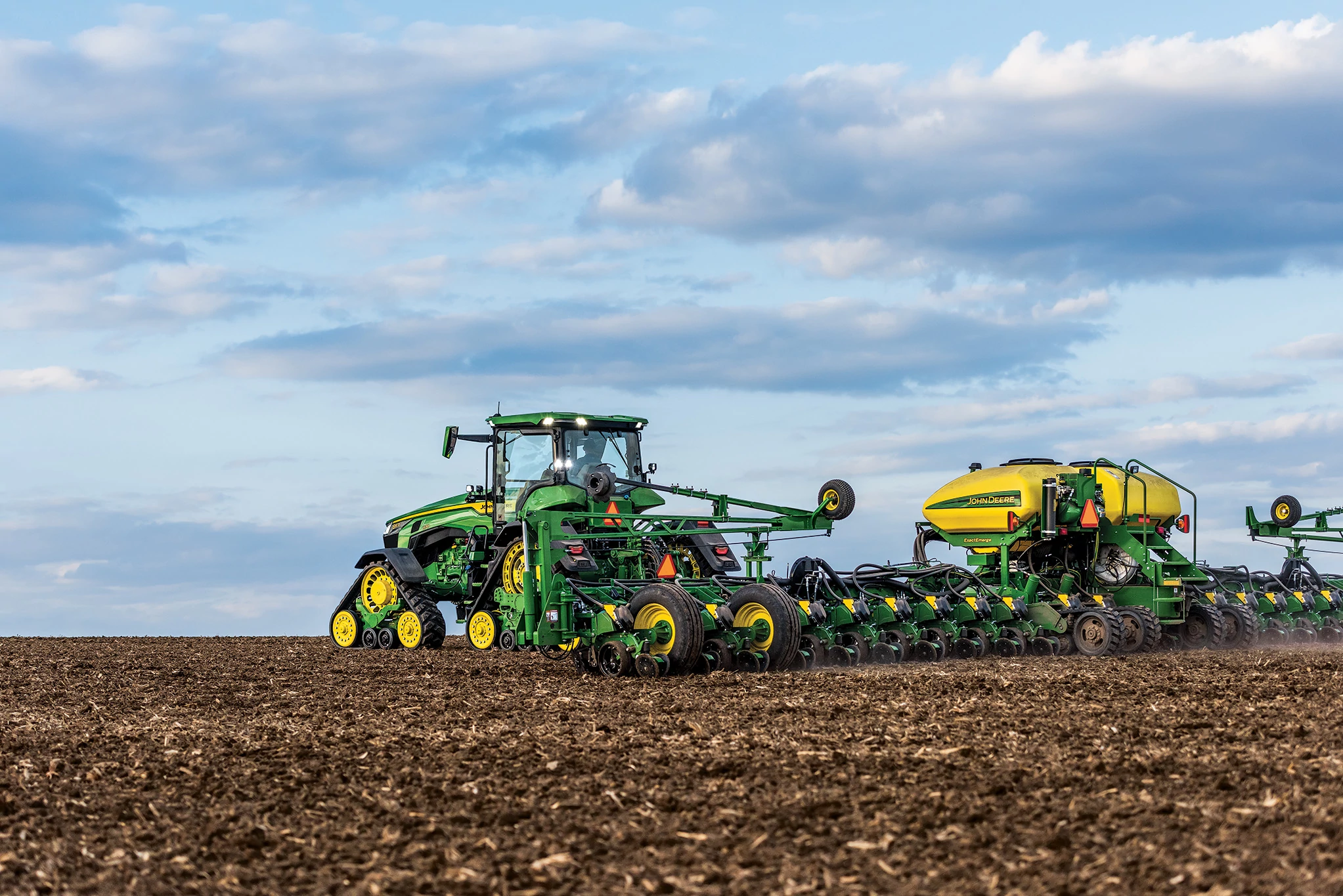 A green John Deere tractor utilizing the JDLink Boost Mini Terminal pulls a large planter across a freshly tilled field beneath a wide sky filled with blue and gray clouds.
