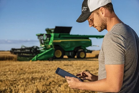 Man in gray shirt and ballcap looking at data on a tablet with a combine operating in the background
