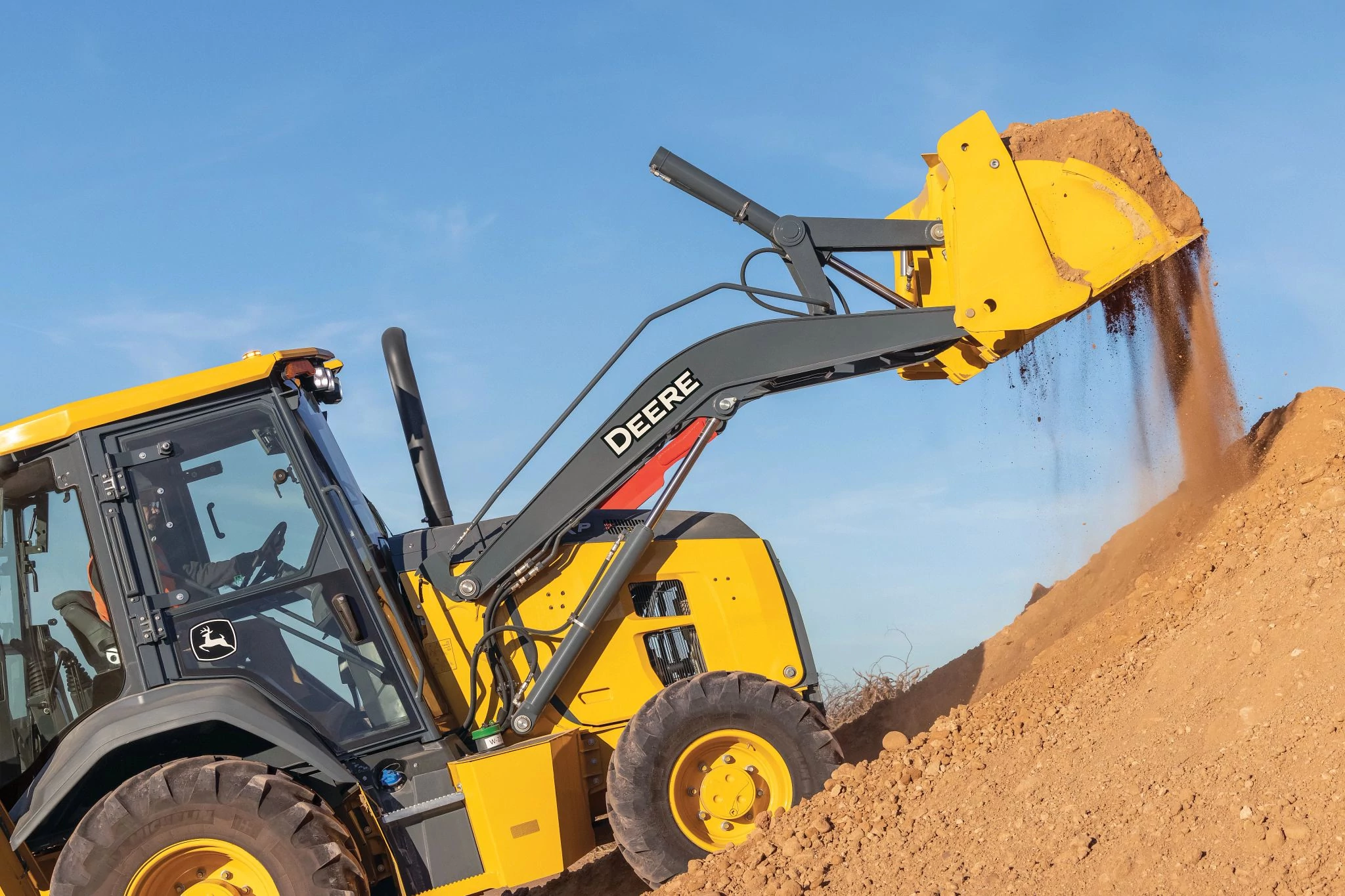 A close-up view of a yellow John Deere backhoe loader lifting a full bucket of sandy soil, with dirt spilling from the raised bucket onto a large mound.