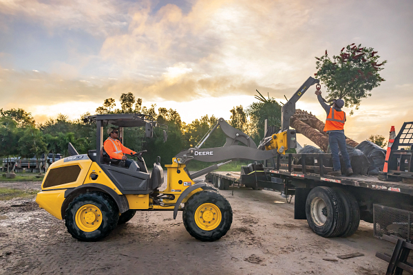 Yellow front-end loader transferring large palm trees to a flatbed truck at a construction site, with two workers in high-visibility vests guiding the load at sunset