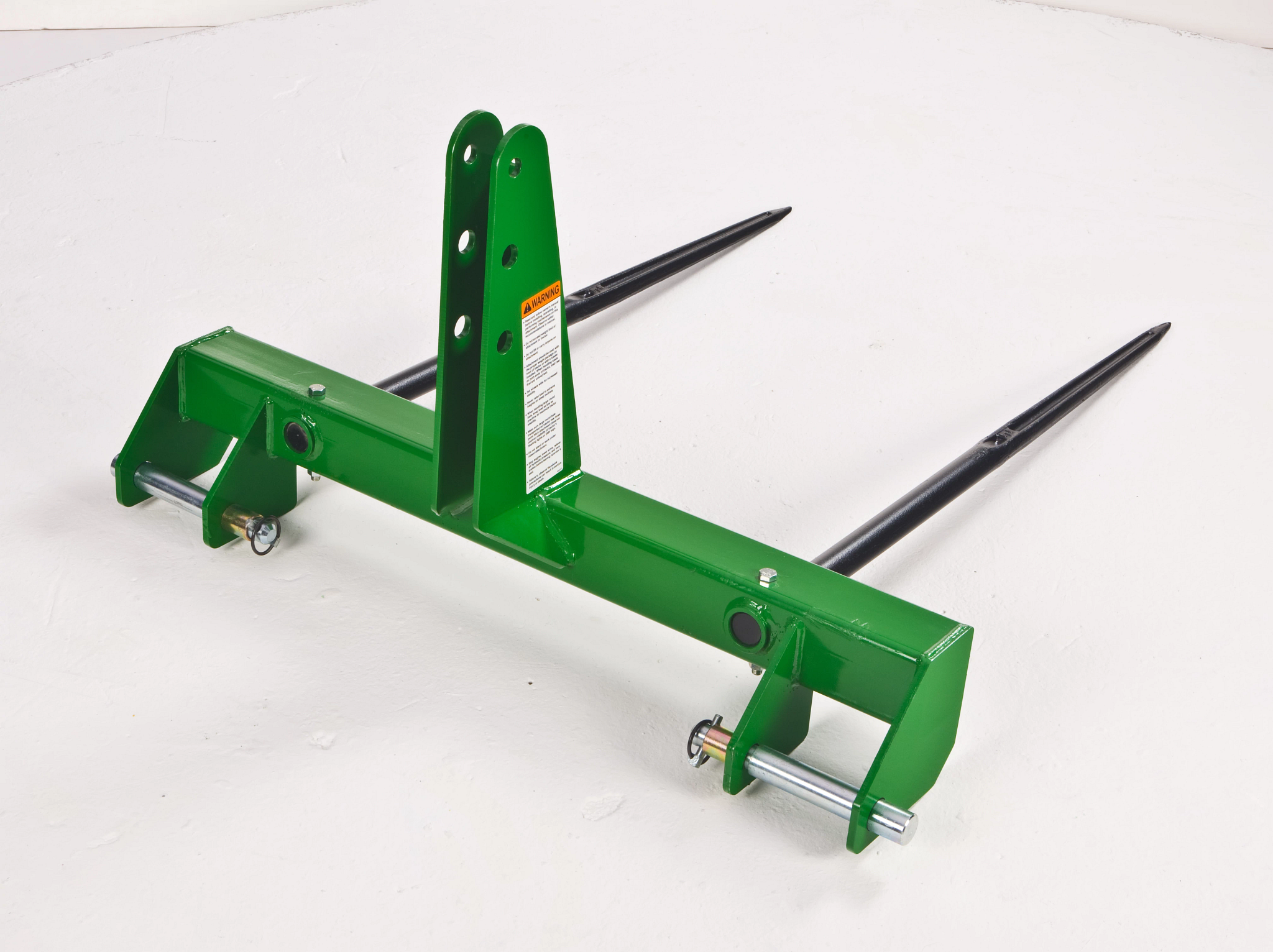 Rear right studio view of a green bale spear resting on a white floor: tubular frame and two pointed spear tines