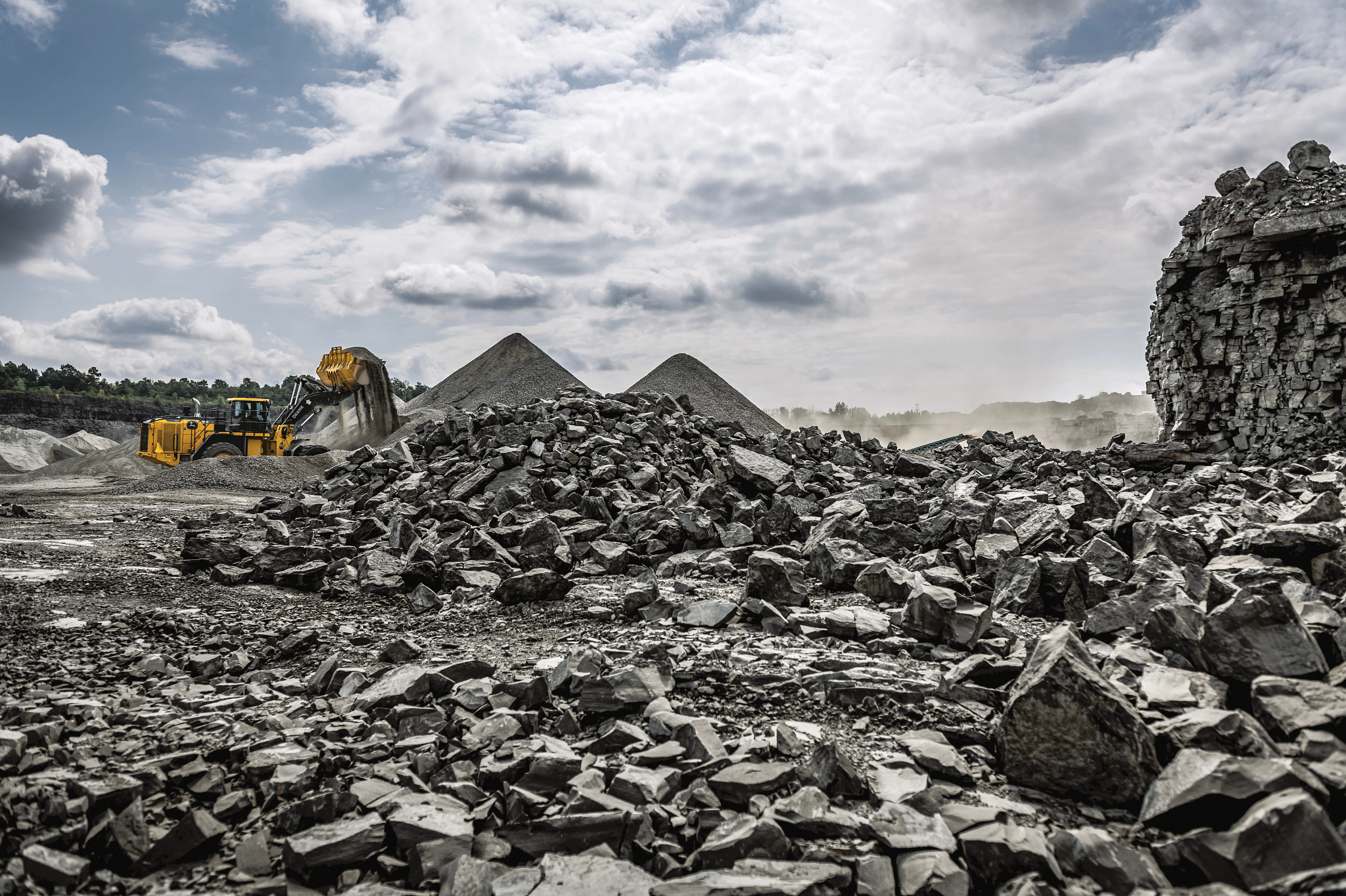 Yellow wheel loader scooping and dumping rock in a quarry, surrounded by large piles of loose stones and gravel under a cloudy sky, with dust rising in the background