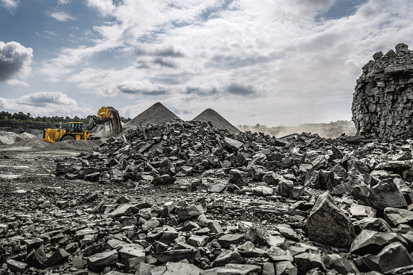 Yellow wheel loader scooping and dumping rock in a quarry, surrounded by large piles of loose stones and gravel under a cloudy sky, with dust rising in the background