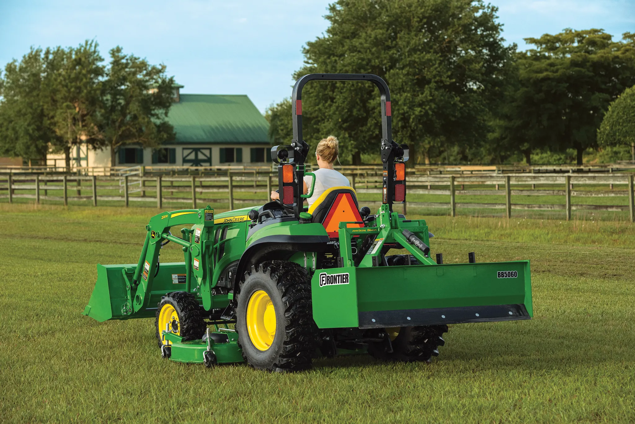 Green compact utility tractor with a front loader and rear implement attachment in a grassy field, surrounded by fencing and trees and farm buildings visible in the background