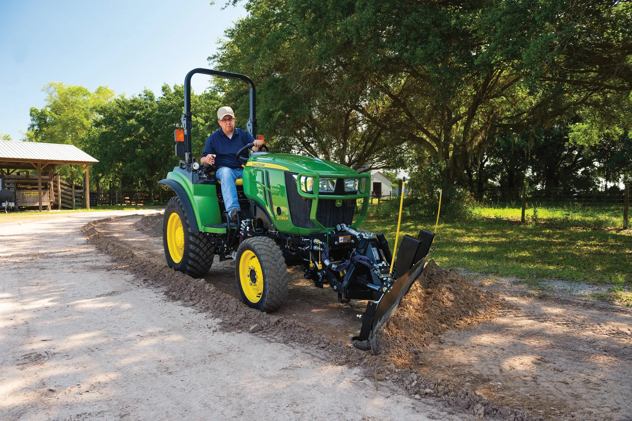 Person operating a compact green tractor with a front attachment leveling a dirt driveway on a rural property with trees, a shed, and dappled sunlight filtering through the foliage
