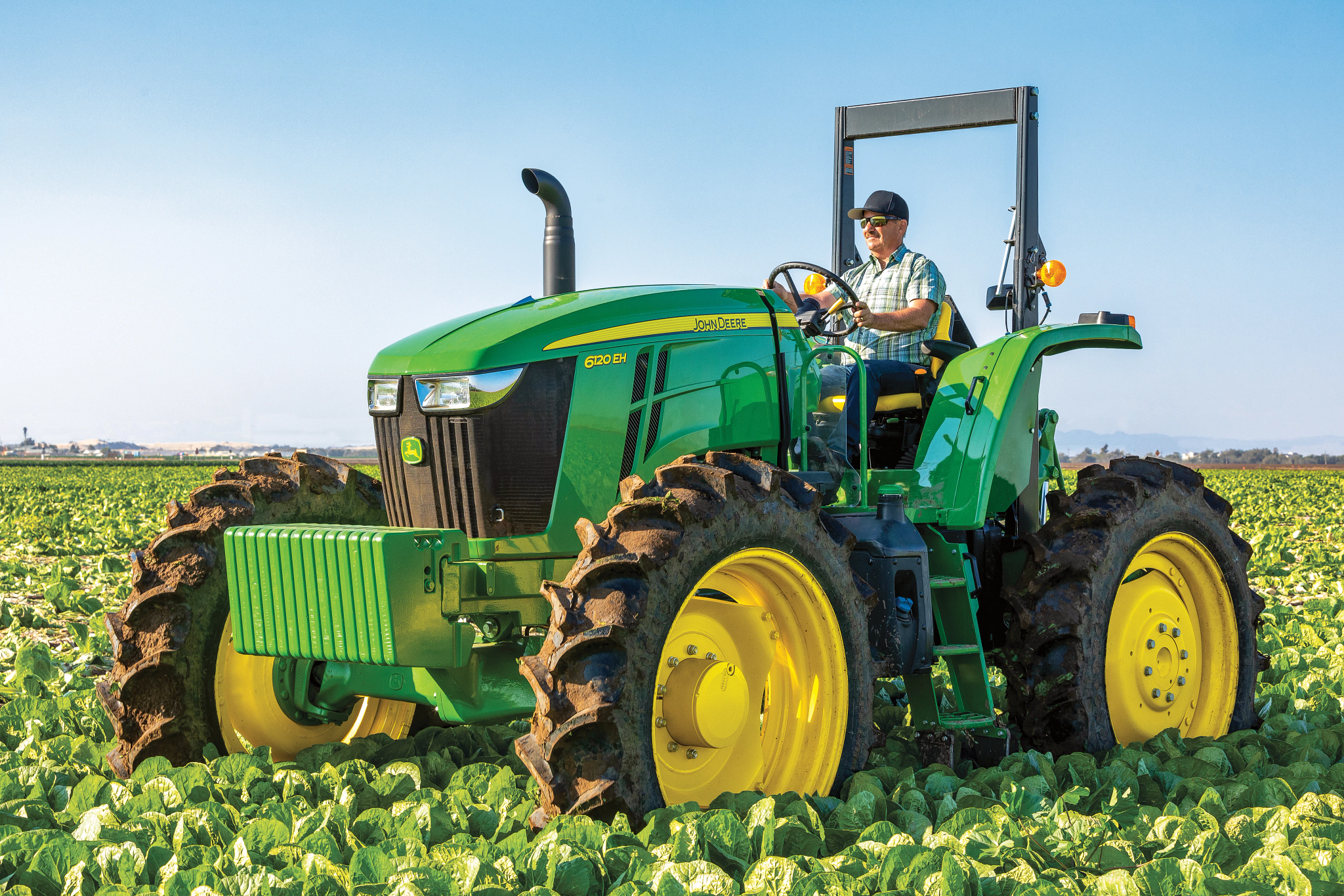 John Deere row‑crop tractor with yellow wheels driving through rows of leafy green crops, fitted with front weights and large rear tires, operating in an open agricultural field under clear blue sky.