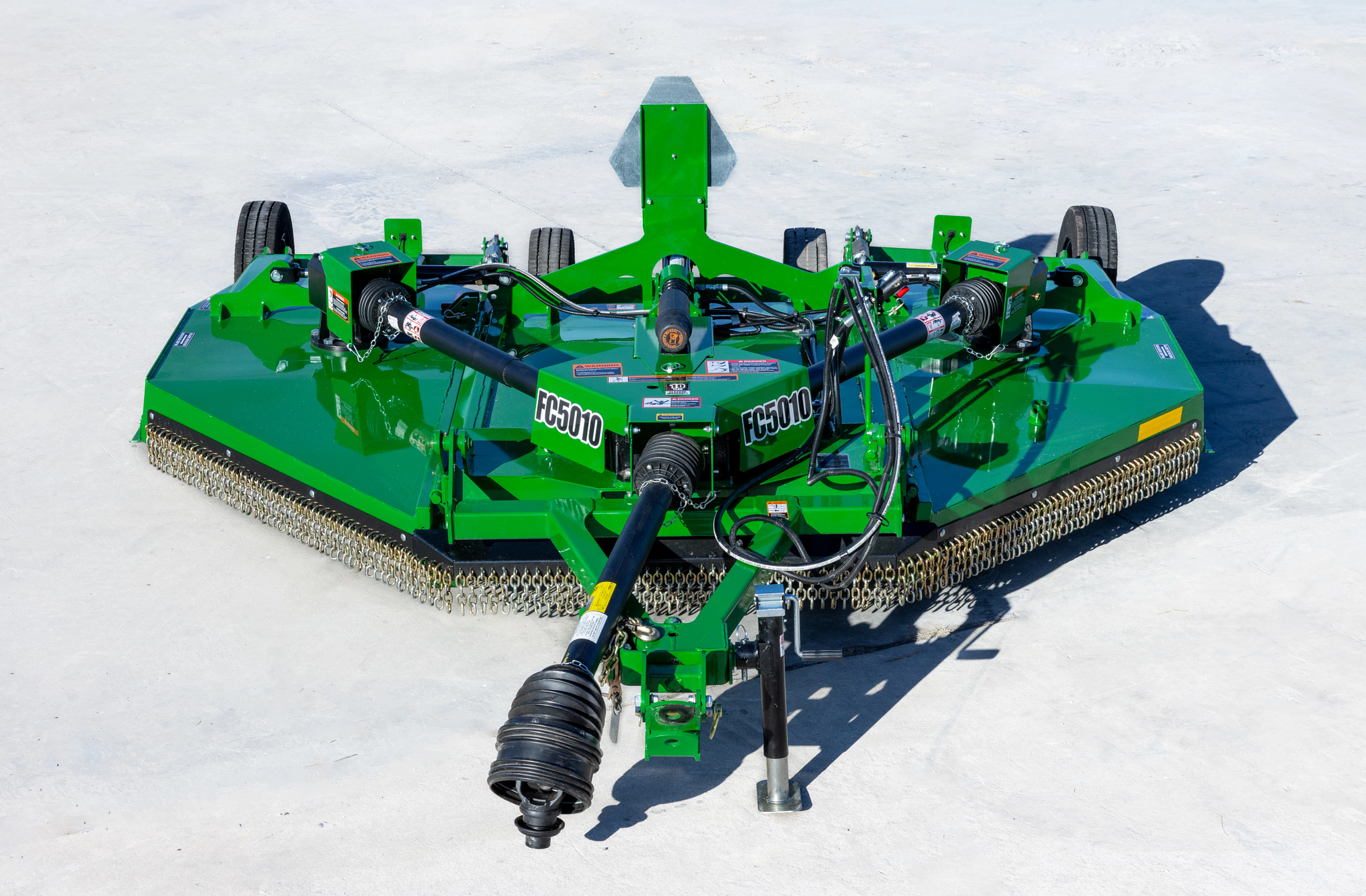 Front view of a green Frontier FC5010 rotary cutter with dual spindles, PTO driveline, hydraulic hoses, transport wheels, and full-width safety chain shielding, positioned on a concrete surface outdoors