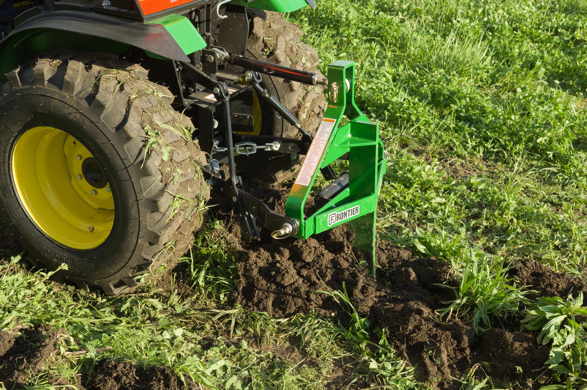 Rear left view of the back of a 2025R Compact Utility Tractor pulling a PS1001 Subsoiler through grassy soil