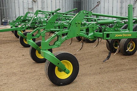 A green chisel plow with wheels parked on a dirt surface, showing its metal frame, shanks, and tines in front of two large metal grain bins