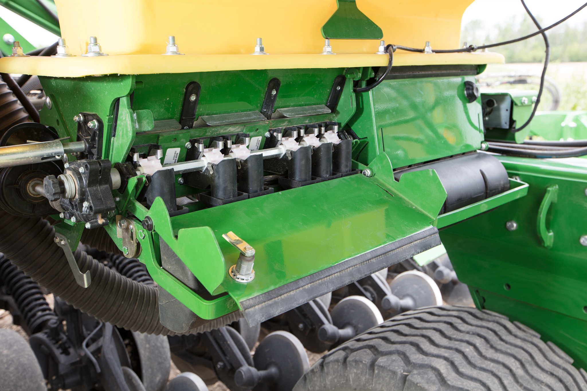 Close-up of machinery showing an open metal housing with seed metering components, drive shaft, hoses, and furrow-opening discs, with a large tire visible on the right