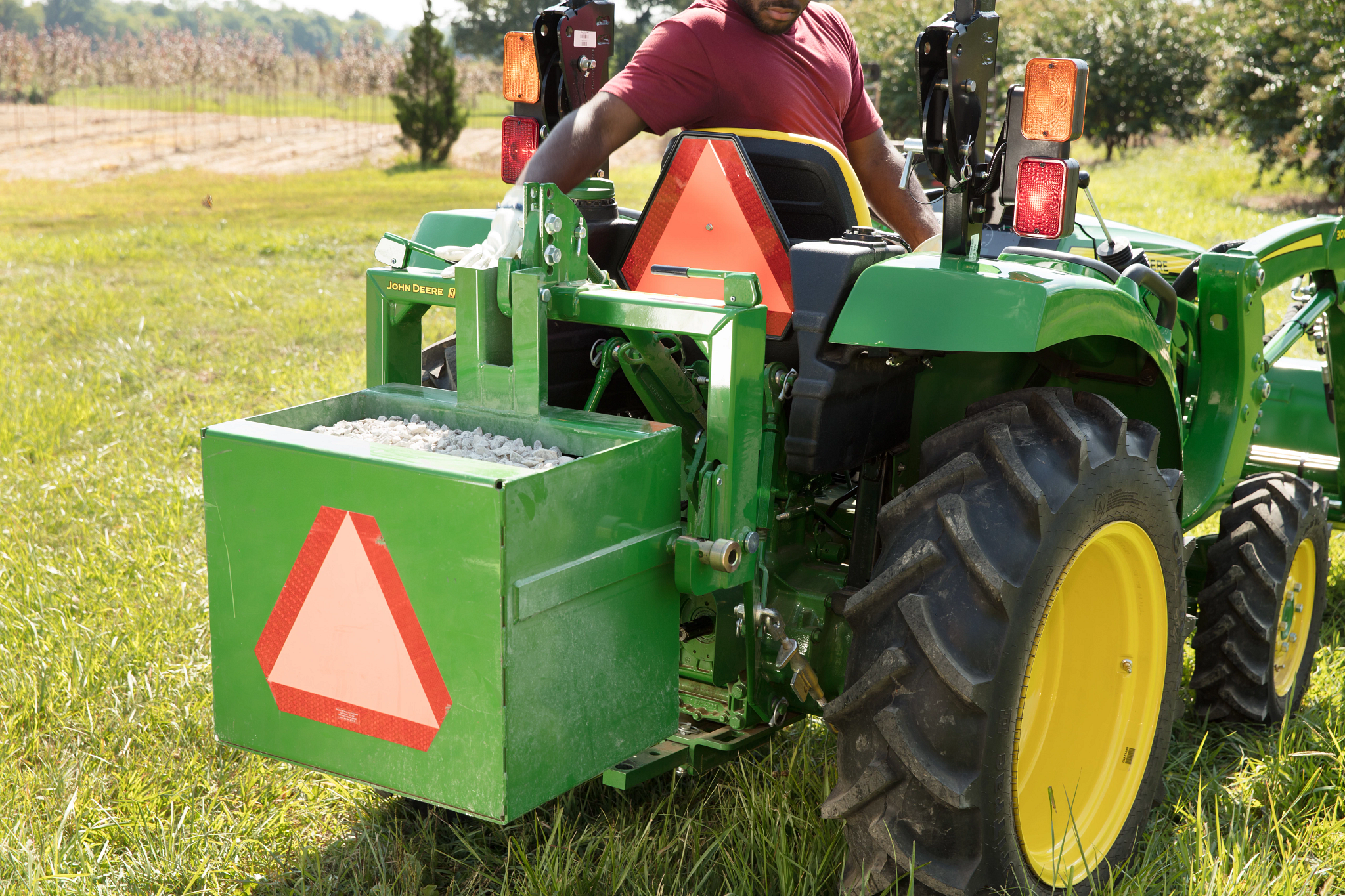 Rear right view of a 3025D Tractor in an orchard with an operator in a red shirt reaching toward the rear box attachment