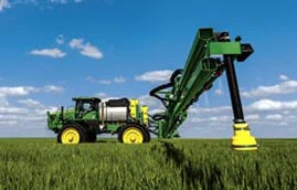 A self‑propelled sprayer in a crop field with the boom extended outward, shown under a blue sky with scattered clouds