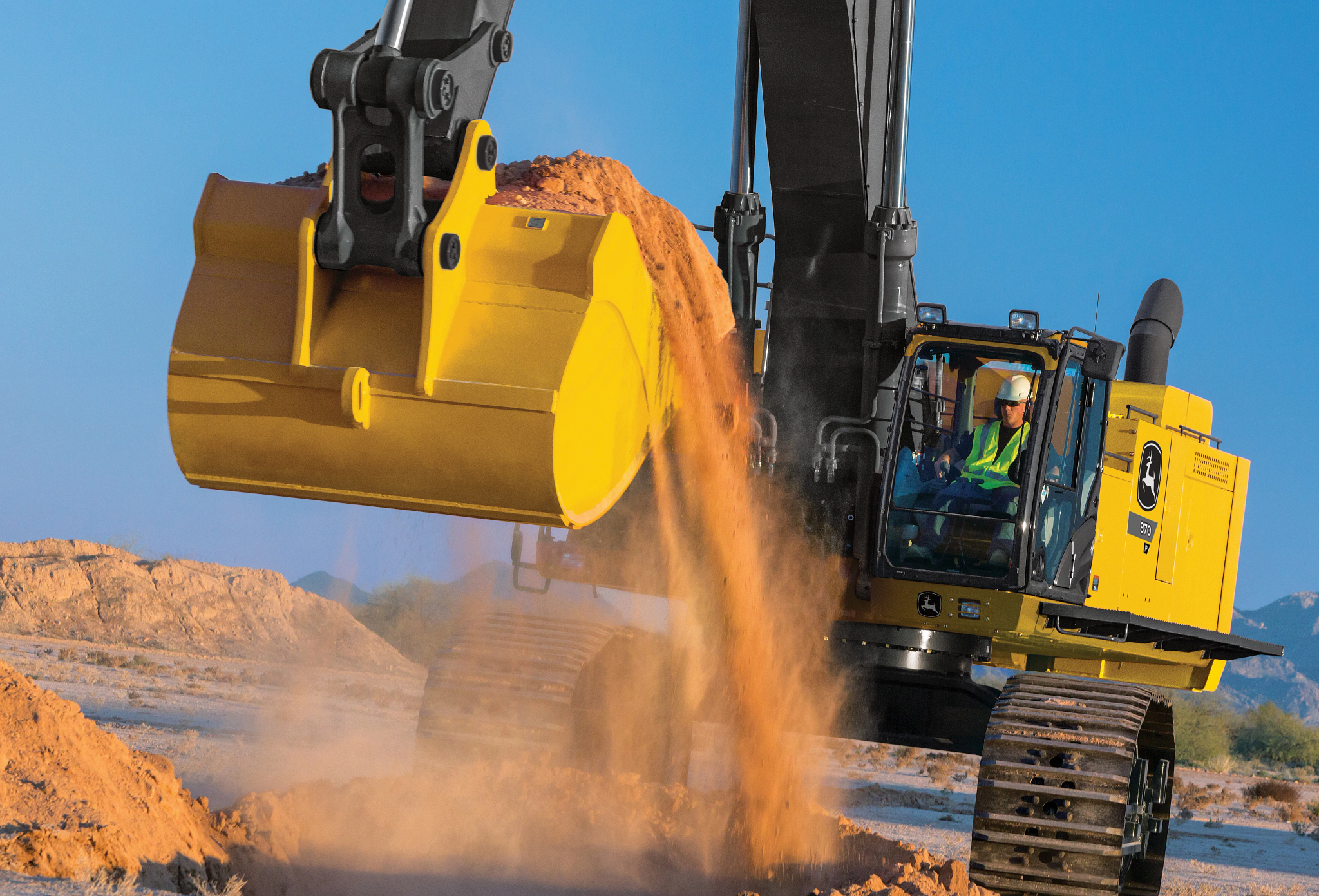 Yellow John Deere excavator with a black arm and large metal bucket releasing a stream of dirt onto the ground at a construction site