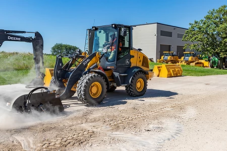 A front-end loader with a brom attachment sweeping a dusty surface outside an industrial building, with other construction equipment parked in the background under a blue sky