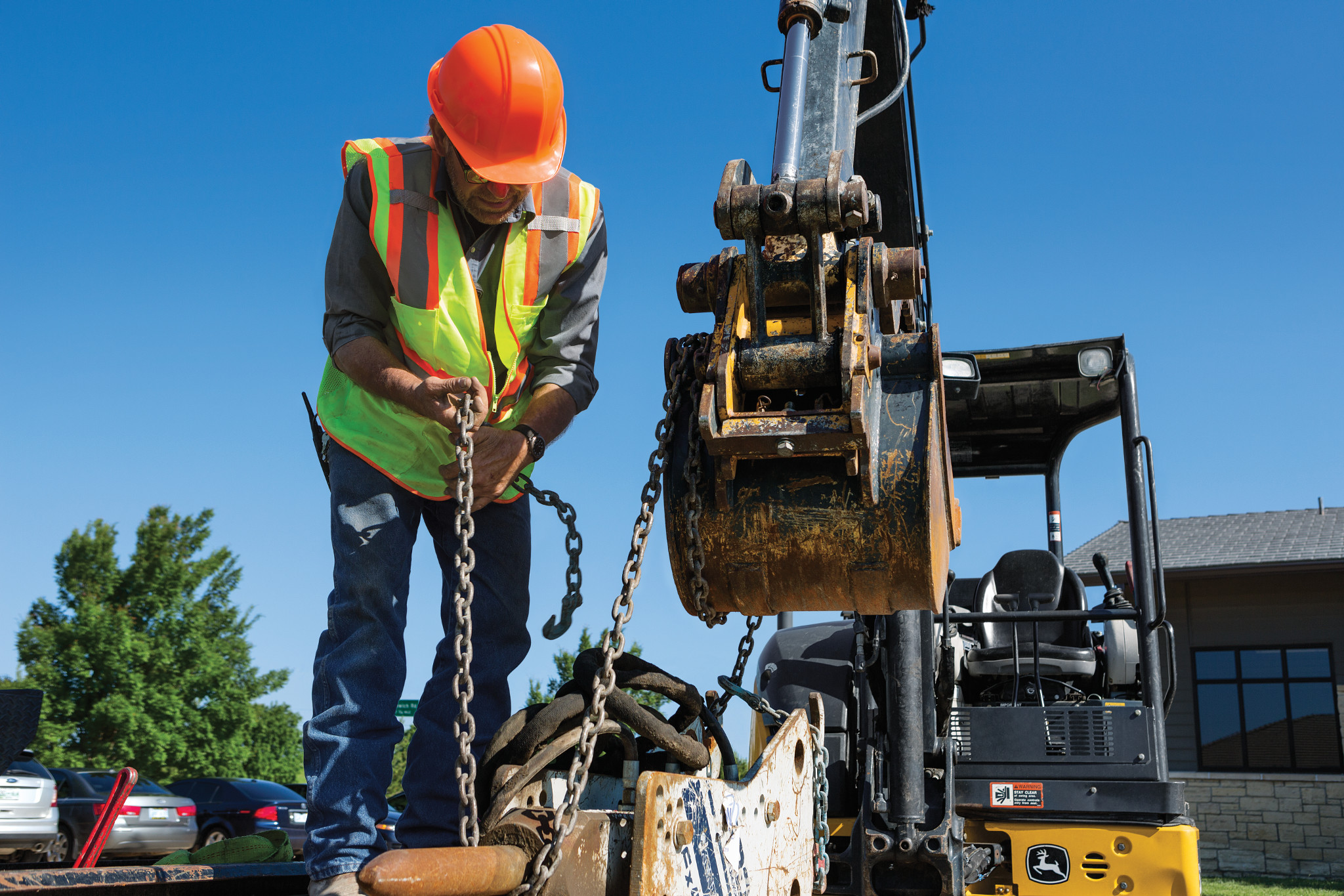 Construction worker in a high-visibility vest and hard hat attaching a chain to equipment while standing next to an excavator bucket at a job site