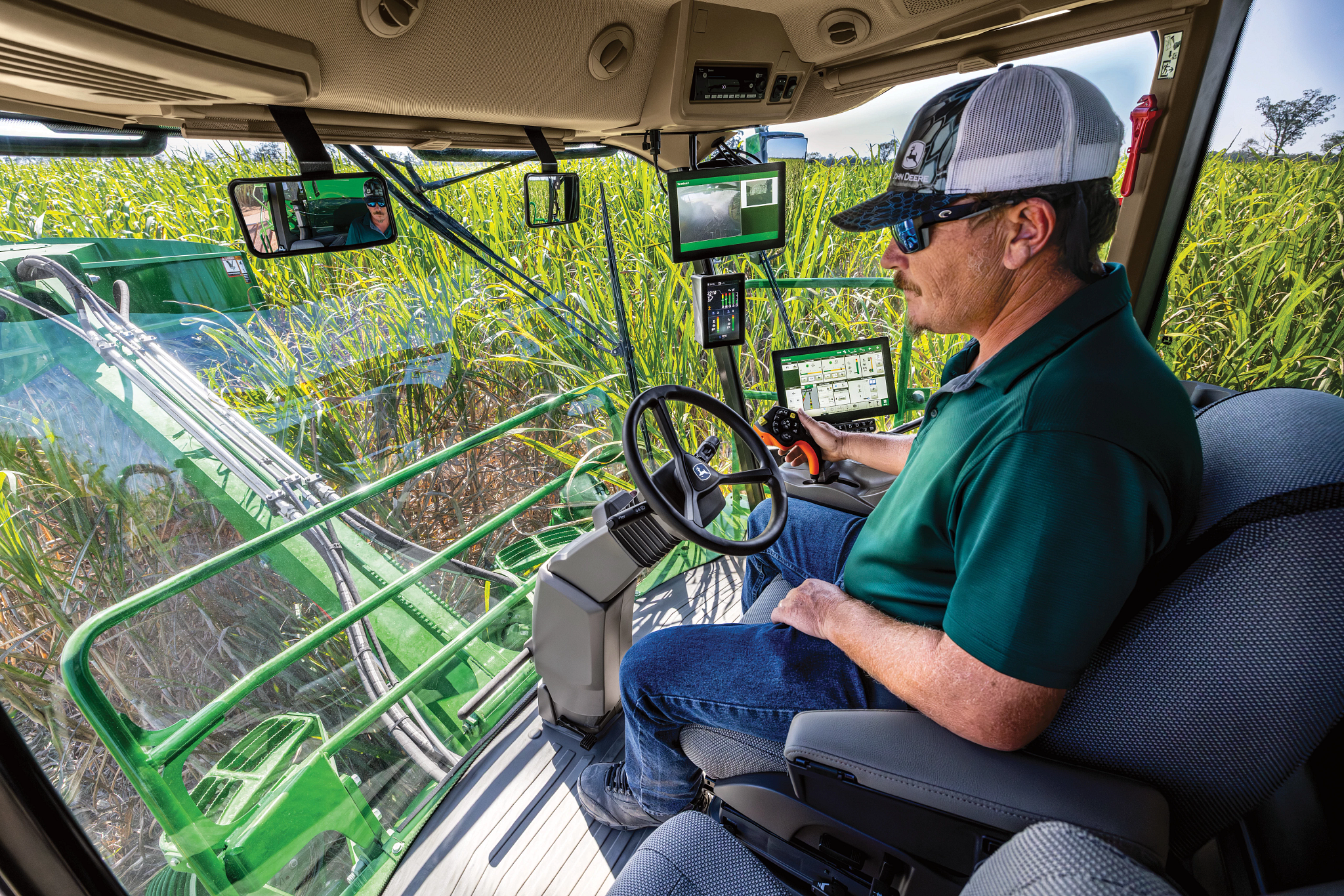 Operator seated inside a farm machine cab, using controls and screens while harvesting sugarcane; green equipment and tall cane visible through the windows under a clear sky