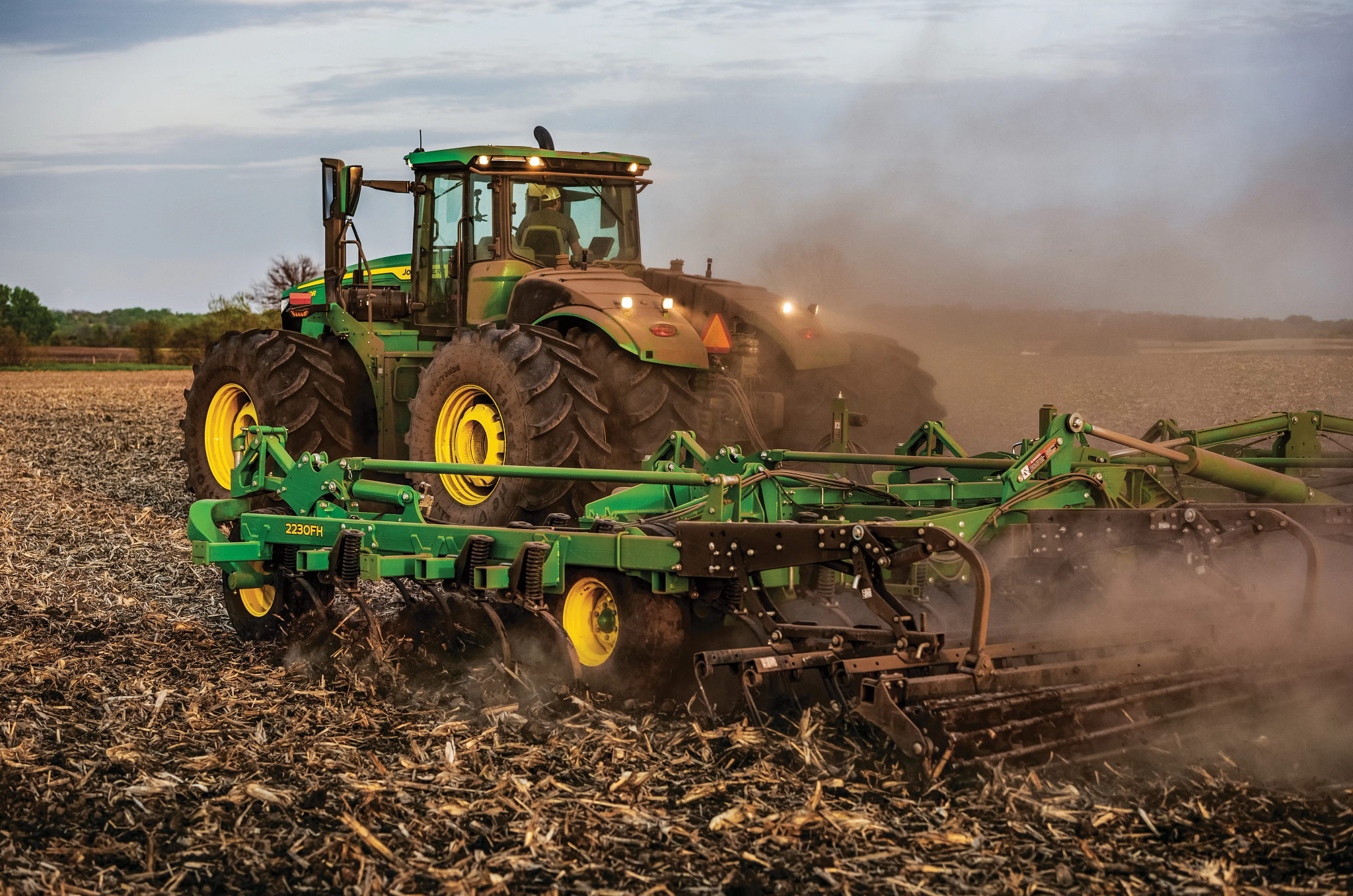 Green tractor with large rear tires towing a wide cultivator across a dry, harvested field with dust rising heavily behind the equipment over flat farmland under a hazy sky