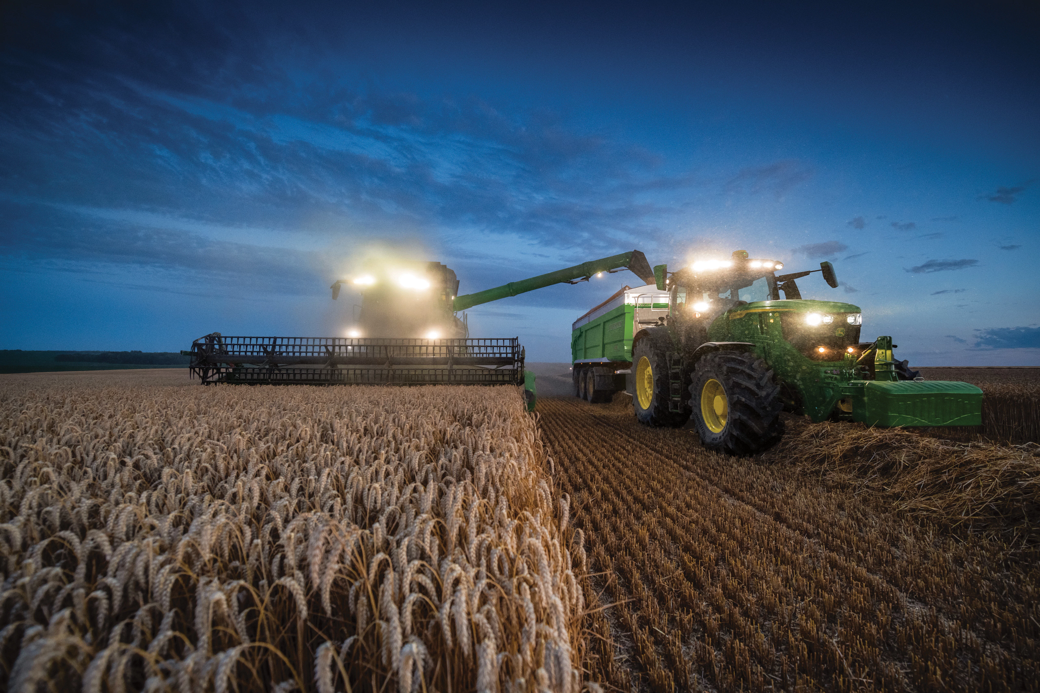 Front view of a T5 700 combine with XA25 header and 6R 250 tractor harvesting a golden field at dusk with their fronts lit up