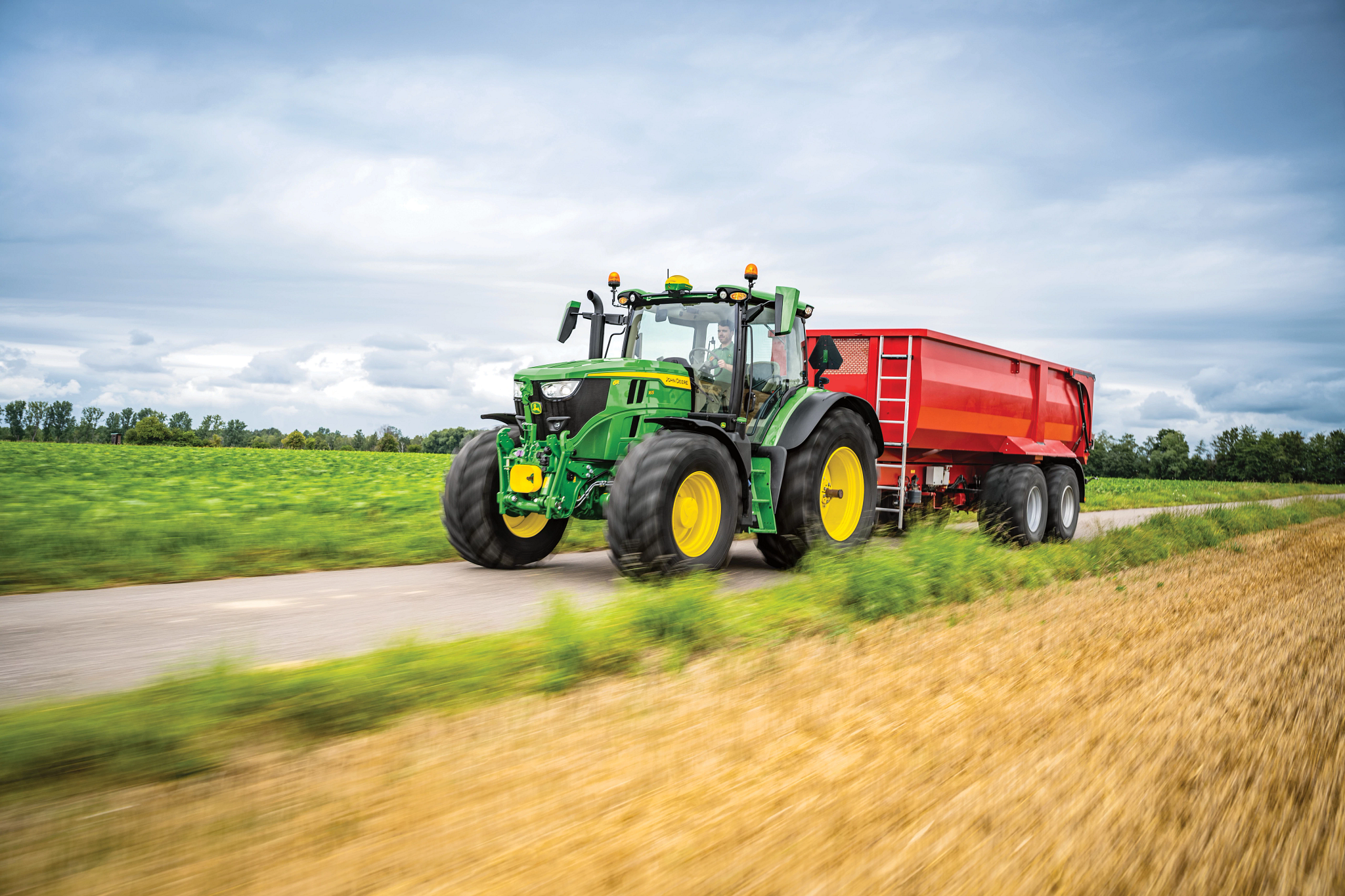 John Deere tractor with yellow wheels towing a red agricultural trailer along a rural paved road, driving past green fields and harvested grass under a cloudy sky.