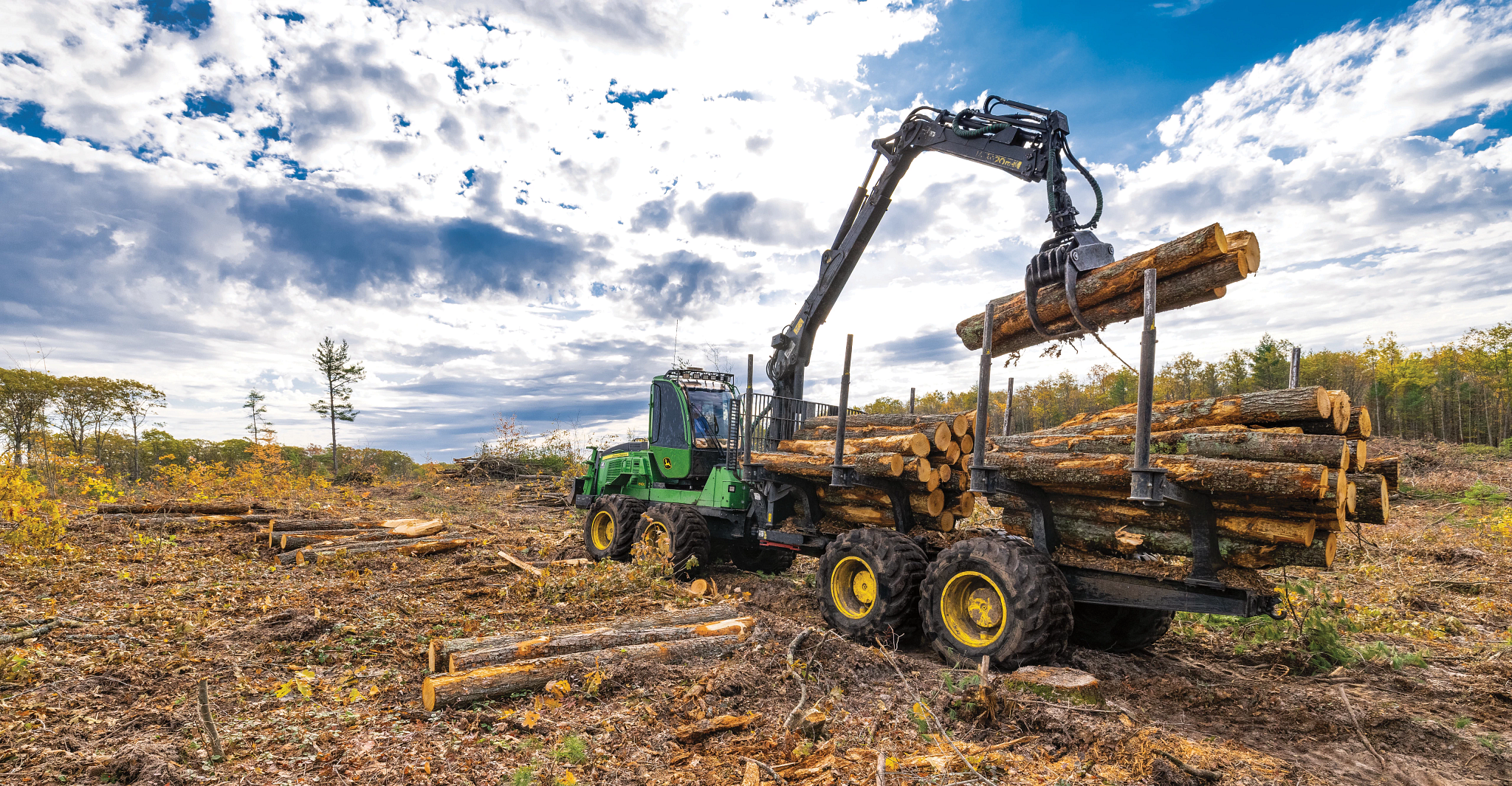 Forestry forwarder loading cut logs onto its trailer using a hydraulic boom and grapple in a harvested timber area under a partly cloudy sky
