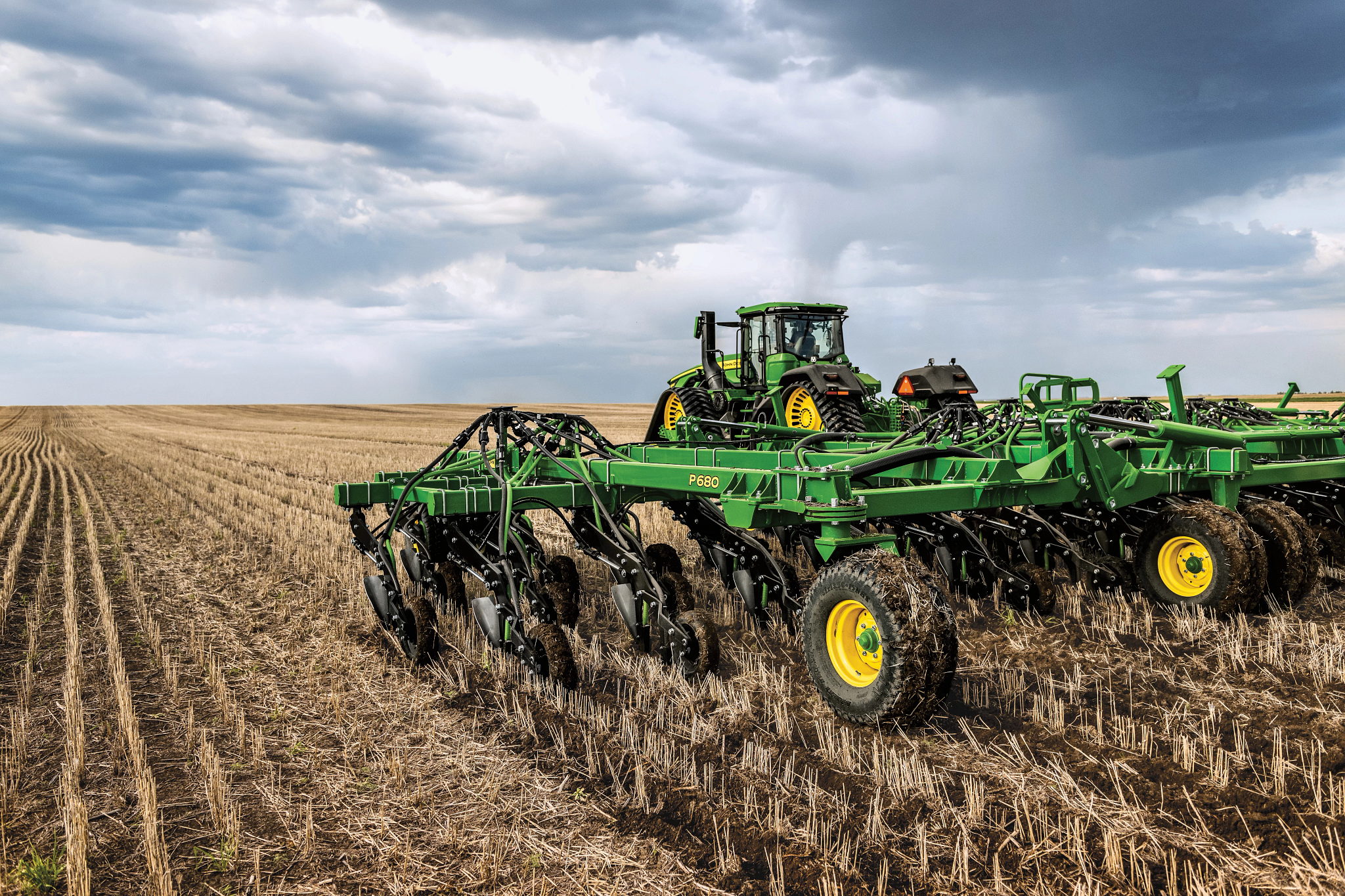 Agricultural tractor pulling a large green tillage implement across a harvested field of dry corn stubble under a cloudy sky, with flat farmland stretching to the horizon
