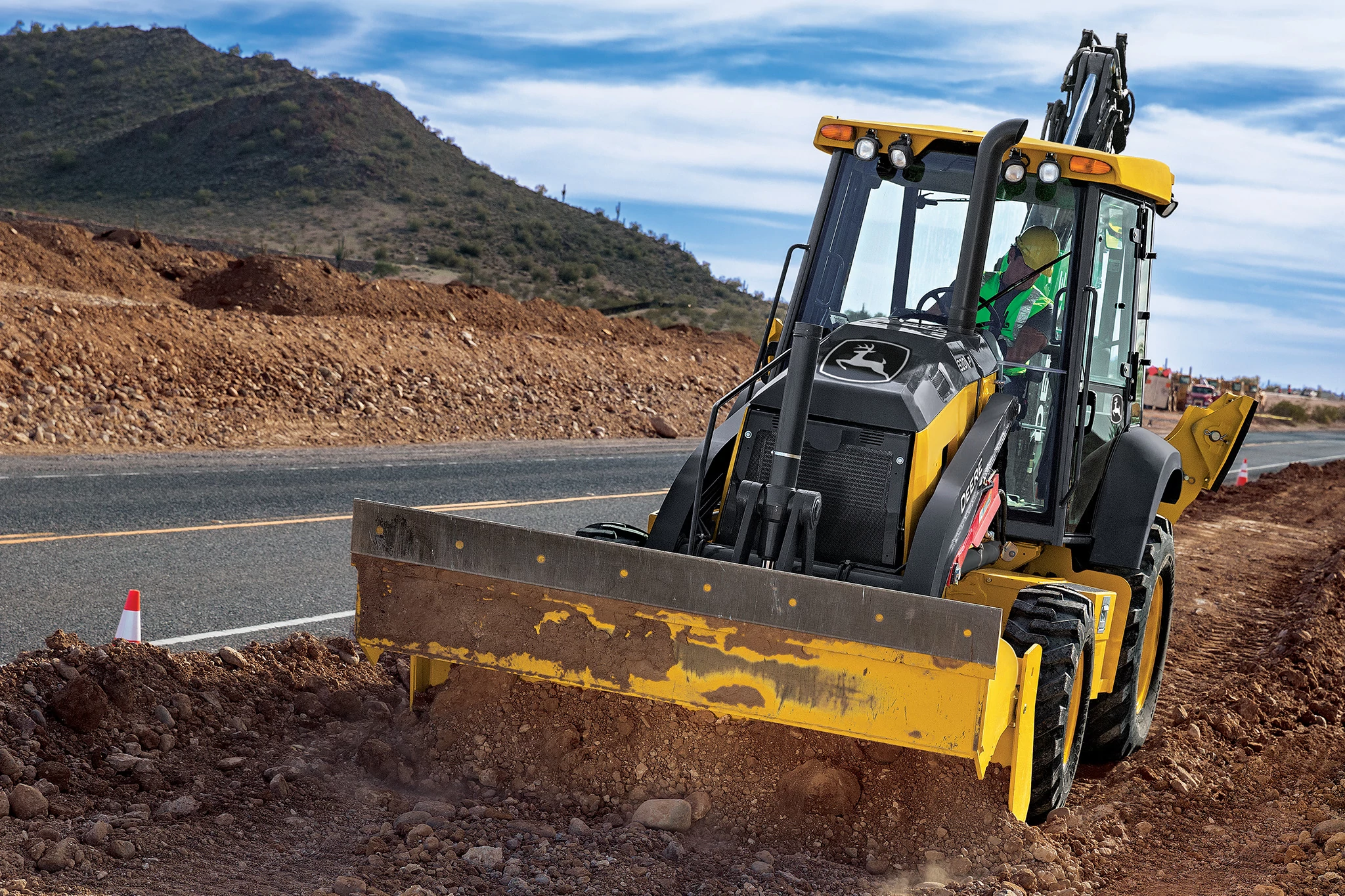 310 G-Tier Backhoe operates along a roadside construction site, pushing soil with a wide front blade in the foreground.