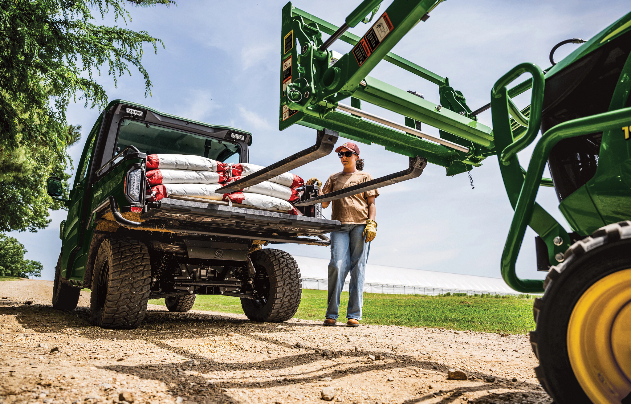 View from a dirt road of a 1025R loader attachment with 120R pallet forks unloading bags from an XUV 845 Gator while an operator looks on from beside it