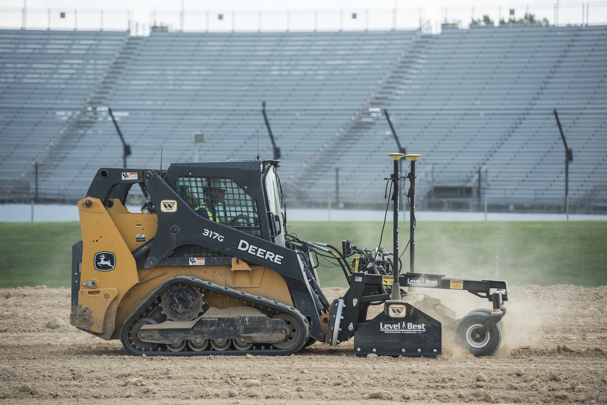 Side right view of a 317G compact track loader with front Level Best attachment working on the dirt infield of a racetrack
