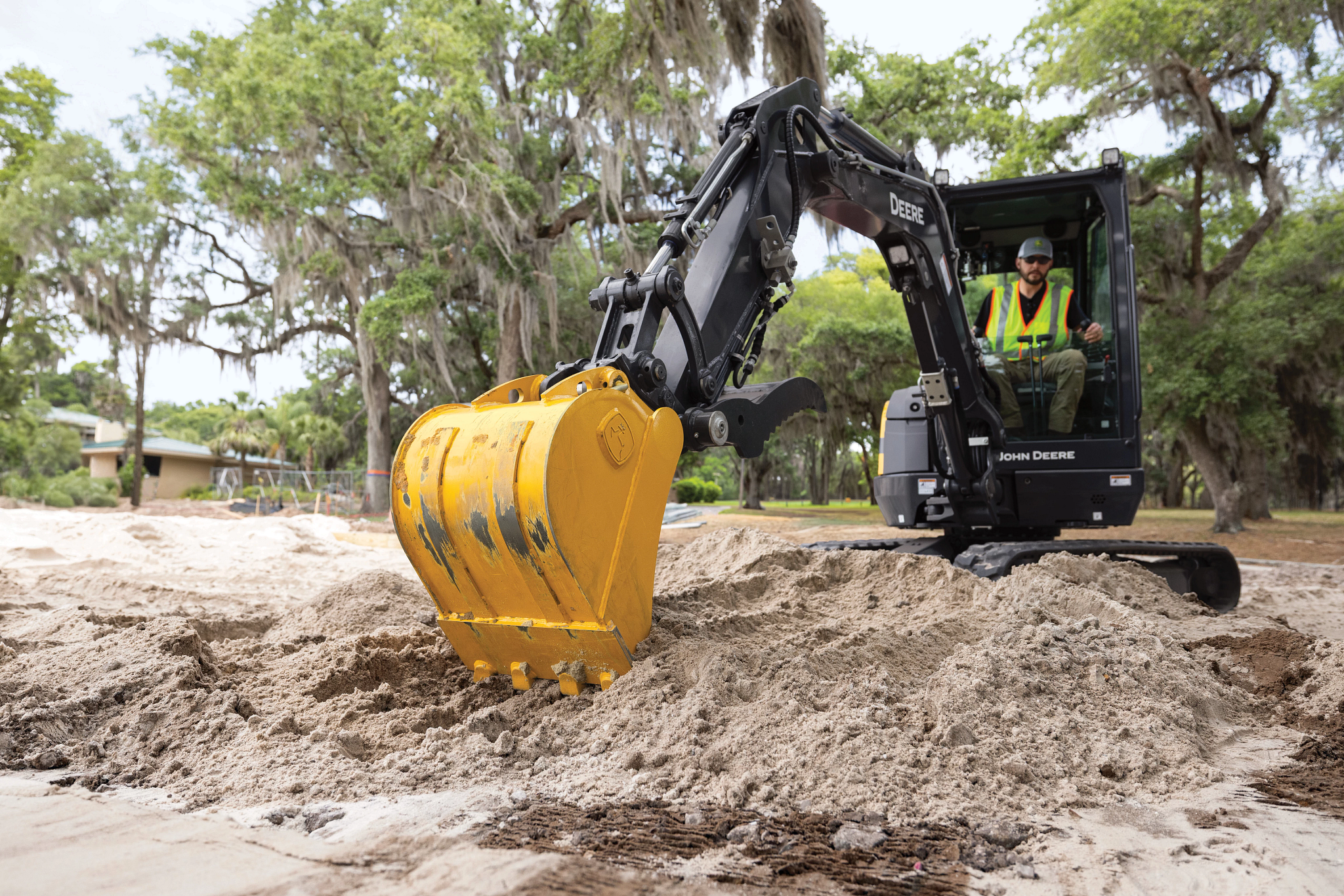Compact excavator operating on a sandy surface, with the bucket lowered into a pile of sand while an operator in a high-visibility vest sits inside the cab, and trees with hanging moss in the background