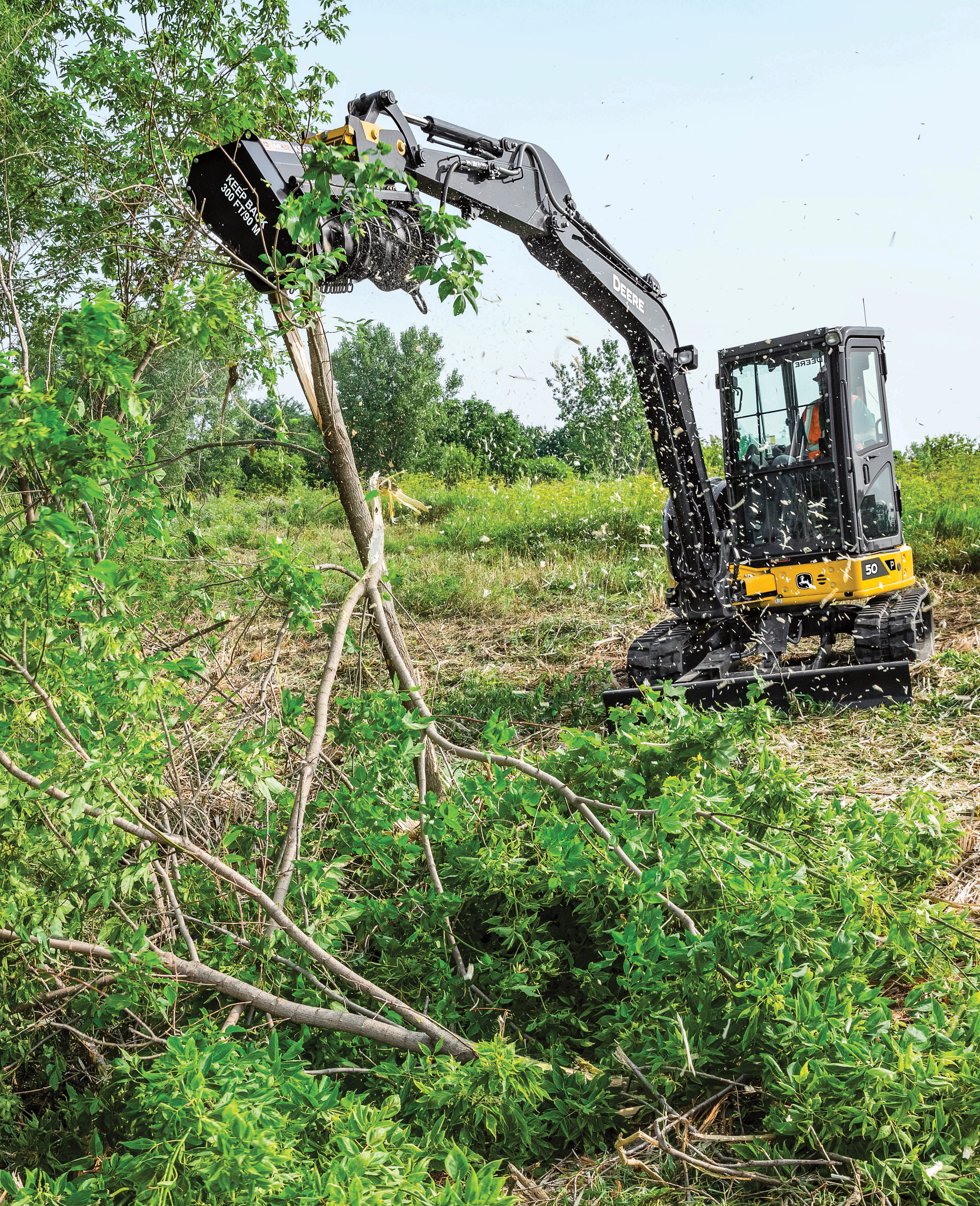 Compact excavator using a mulching head to cut down and clear small trees and brush in a grassy field, with debris falling and dense greenery in the foreground