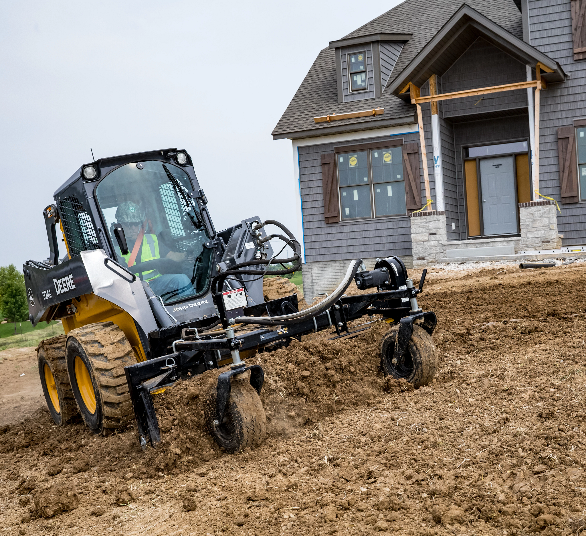 Front right view of a 324G skid steer using attached PR72B angle power rake in the soil in front of a new-build house