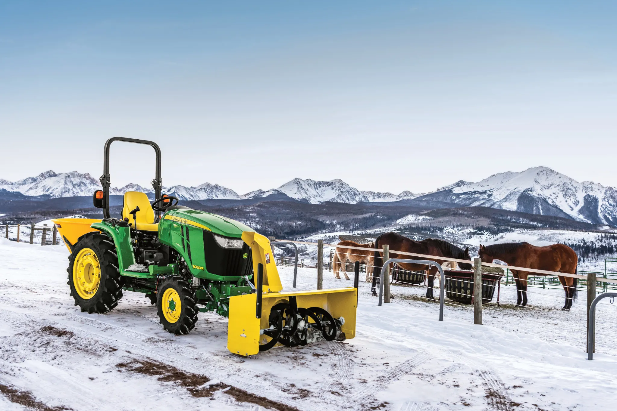 Front right view of an OOS 3039R compact utility tractor parked on a snowy road with horses and mountains in the background and yellow front-attached 60 inch snow blower