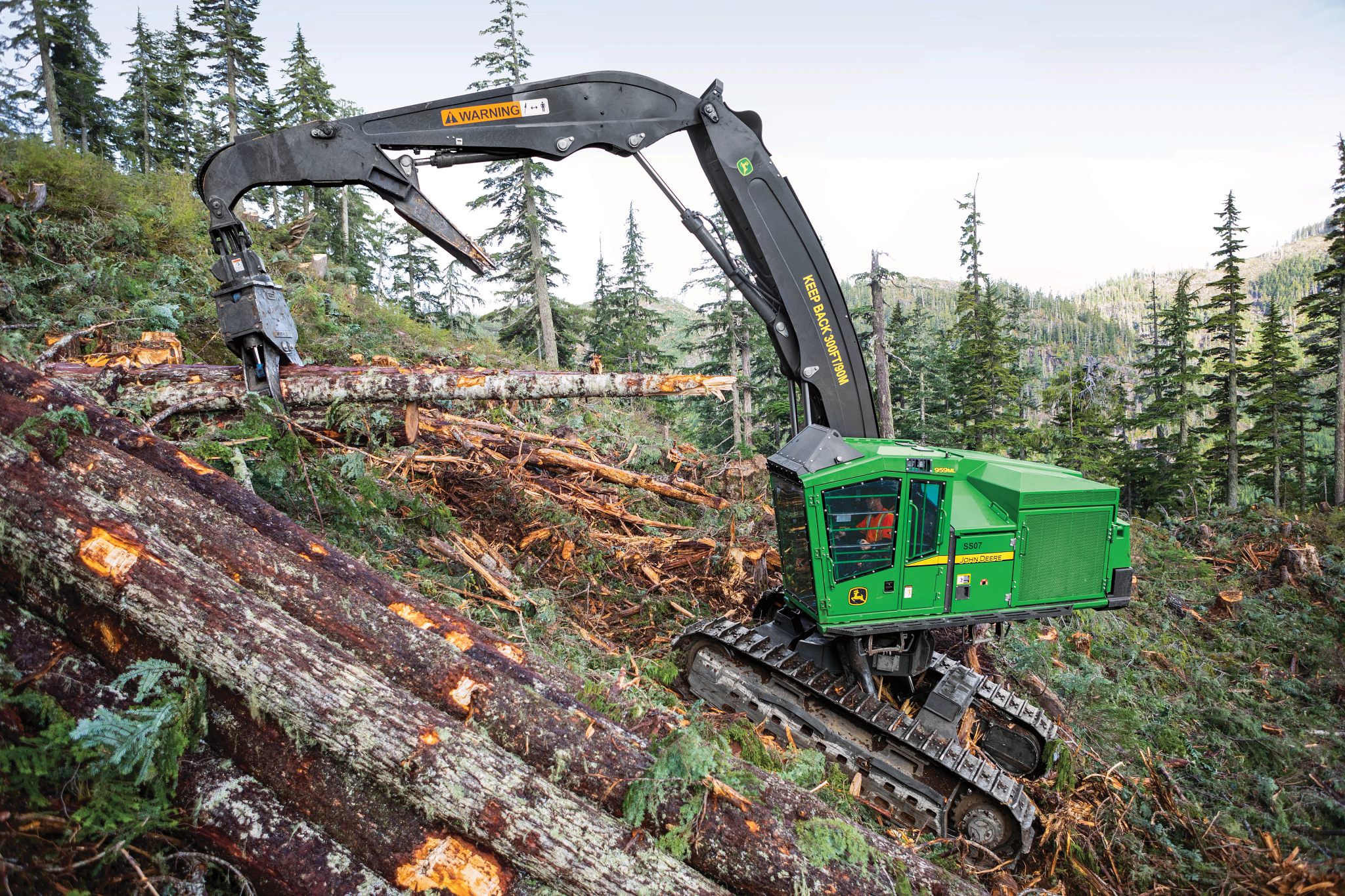 Tracked forestry machine on a steep, wooded slope using a grapple attachment to handle fallen logs, surrounded by trees and cut timber