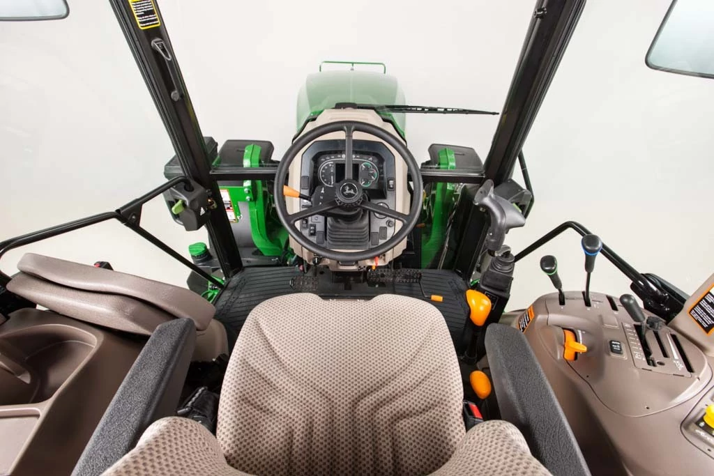 Operator’s view inside John Deere tractor cab showing fabric seat, steering wheel with instrument cluster, right‑hand console with levers and controls, and wide windshield on a clean studio background.