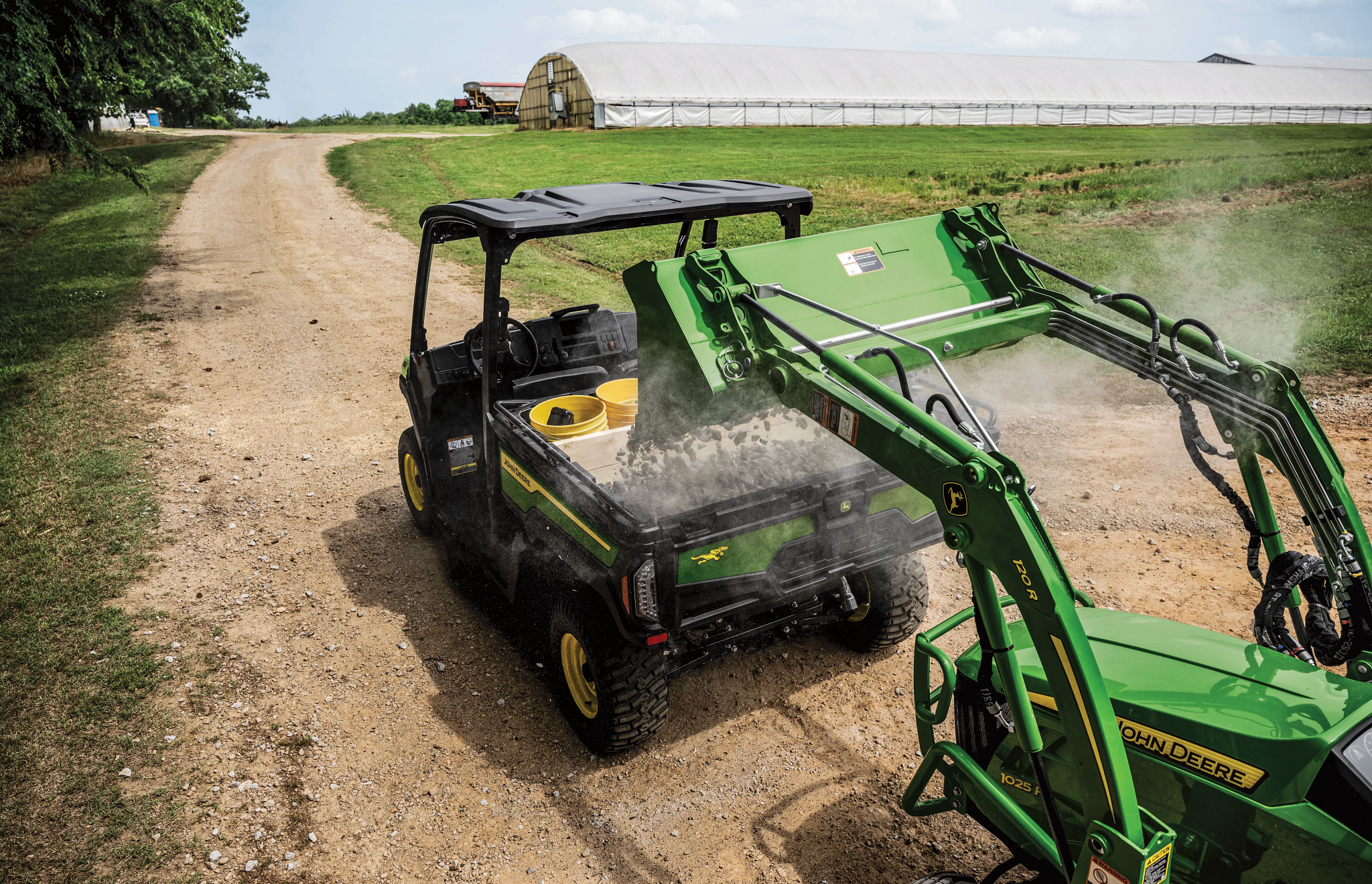 Top-down angled view of an XUV 845E Gator with two yellow buckets in the cargo box being loaded with gravel from behind via green 120R loader attachment on a compact utility tractor