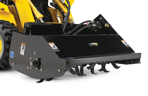 Close-up of a rotary tiller attachment with a black metal housing and curved tines along the bottom, connected to the hydraulic arms and hoses of a skid steer