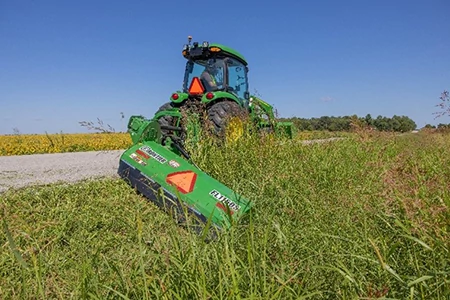 Rear view of tractor pulling a mower attachment through tall grass along a gravel path, with cut vegetation visible in the foreground.