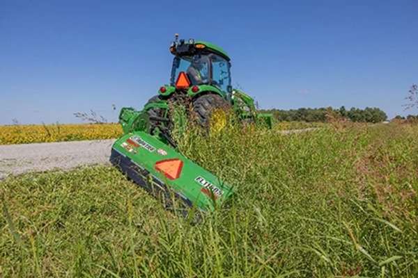 Rear view of tractor pulling a mower attachment through tall grass along a gravel path, with cut vegetation visible in the foreground.