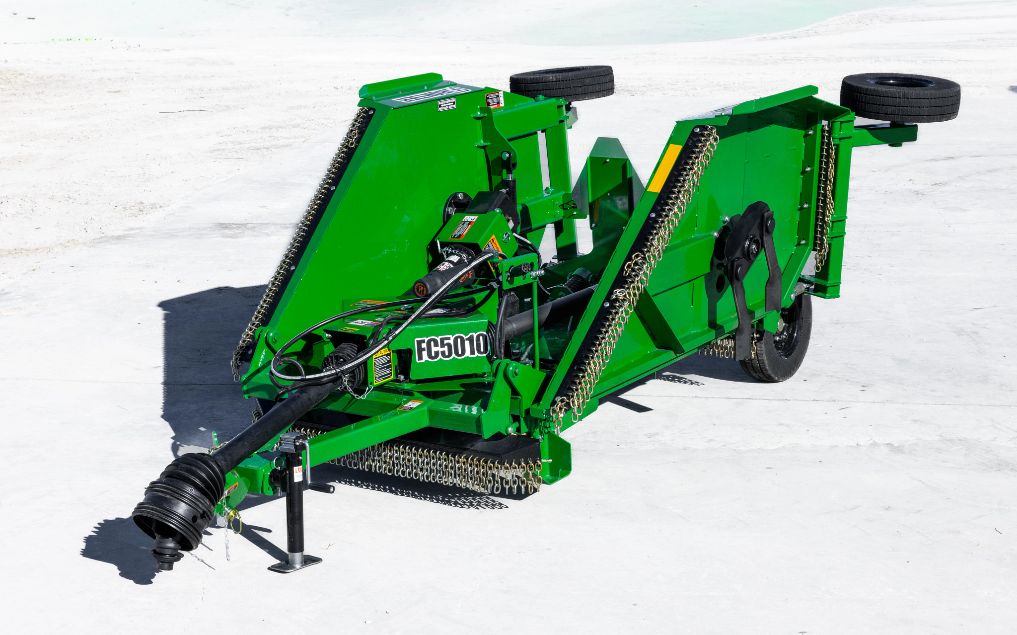 Front three-quarter view of a green Frontier FC5010 rotary cutter in folded transport position, showing raised wing decks, PTO driveline, hydraulic hoses, transport wheels, and safety chain shielding on a concrete surface