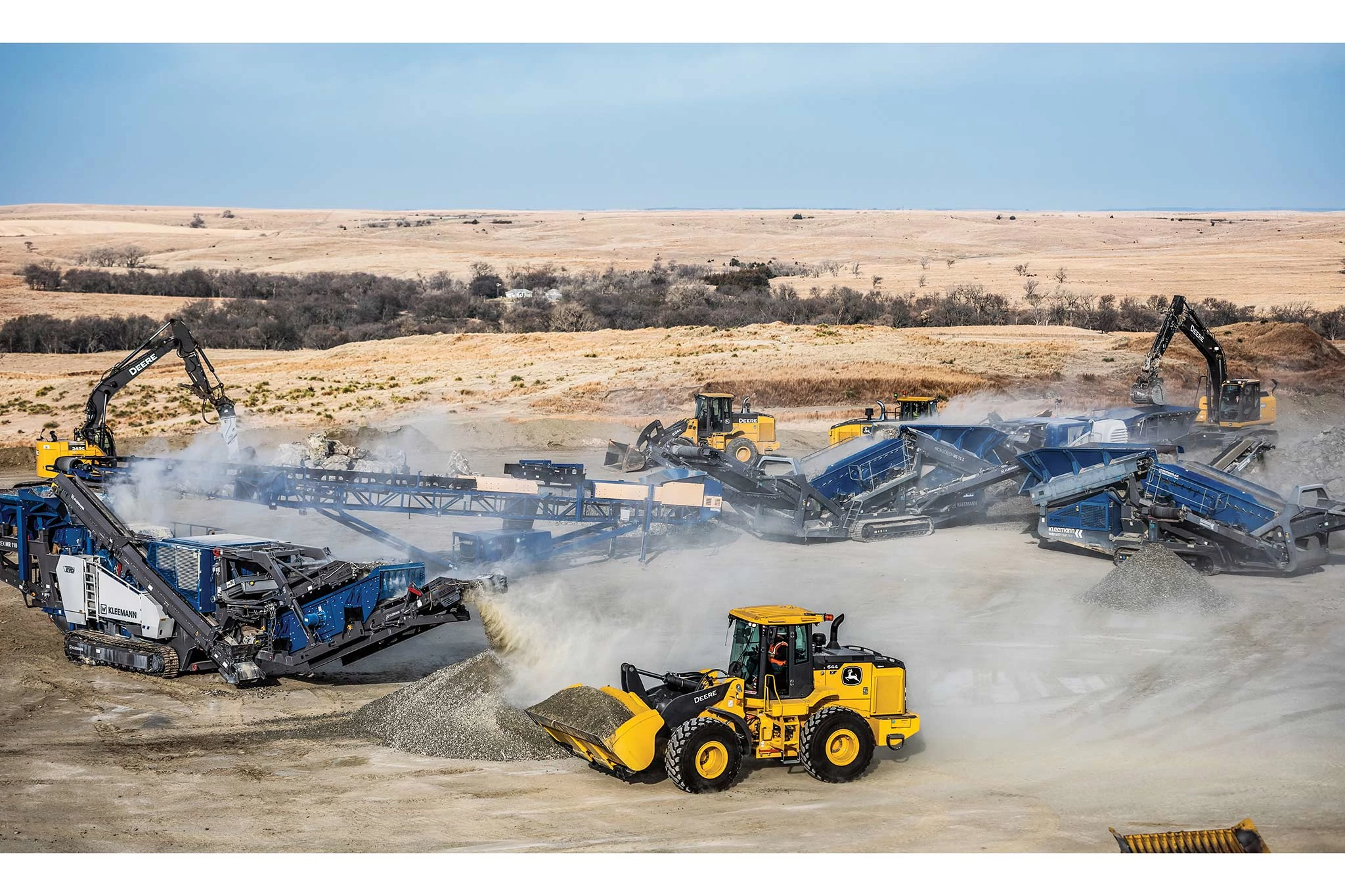 Front aerial view of a 644 X-Tier wheel loader near a gravel pile in front of blue rock crushers and yellow excavators in a quarry