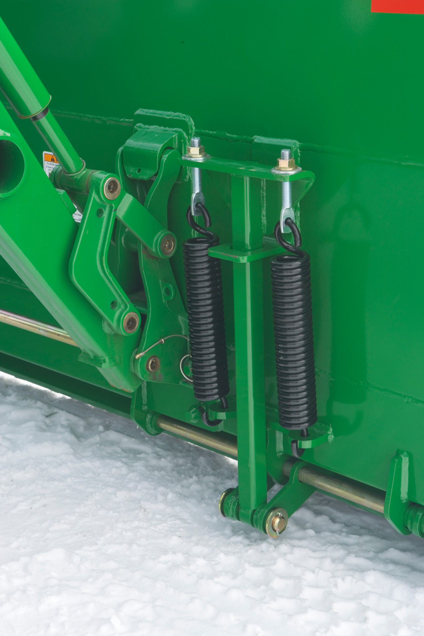 Close-up of a snow push frame showing two vertical tension springs mounted on brackets above a metal bar, positioned over snow-covered ground