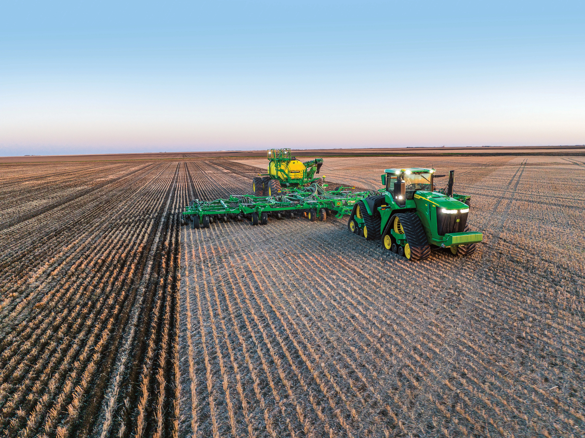 Tractor with dual tracks pulling a large green air seeder and planter across a wide, flat field of evenly spaced crop rows, viewed from above under a clear sky with a distant horizon.