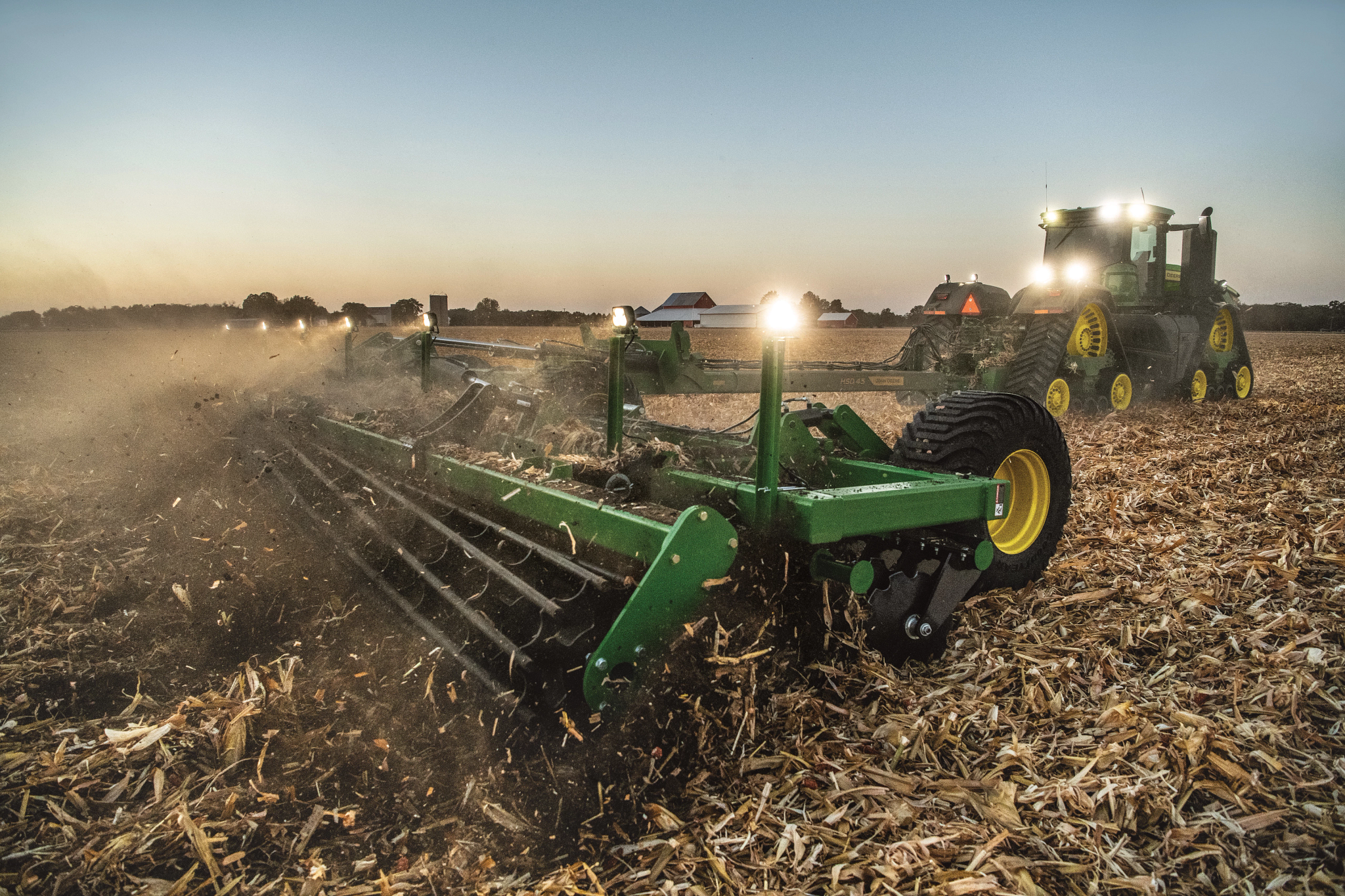 Green tractor pulling a large tillage implement across a dry, harvested field at sunset, kicking up dust and crop residue under a hazy sky with work lights on both the tractor and implement